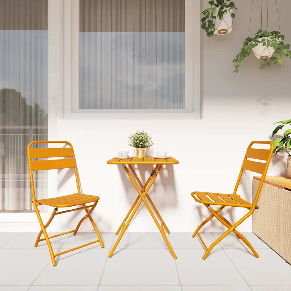 Una mesa amarilla y dos sillas en un patio. Una pequeña planta y dos tazas están sobre la mesa. Dos plantas colgantes arriba.