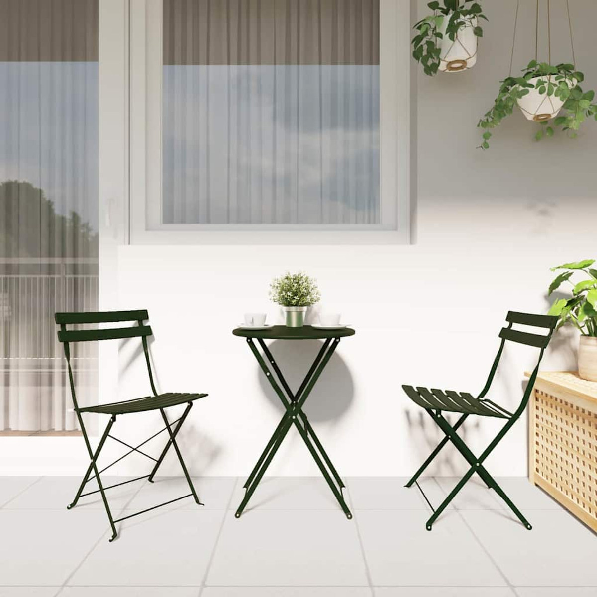 Dunkelgrünes Bistro-Set auf einer Terrasse mit zwei Stühlen und einem kleinen Tisch. Ein Fenster, Pflanzen und weiße Wände vervollständigen die Szene.