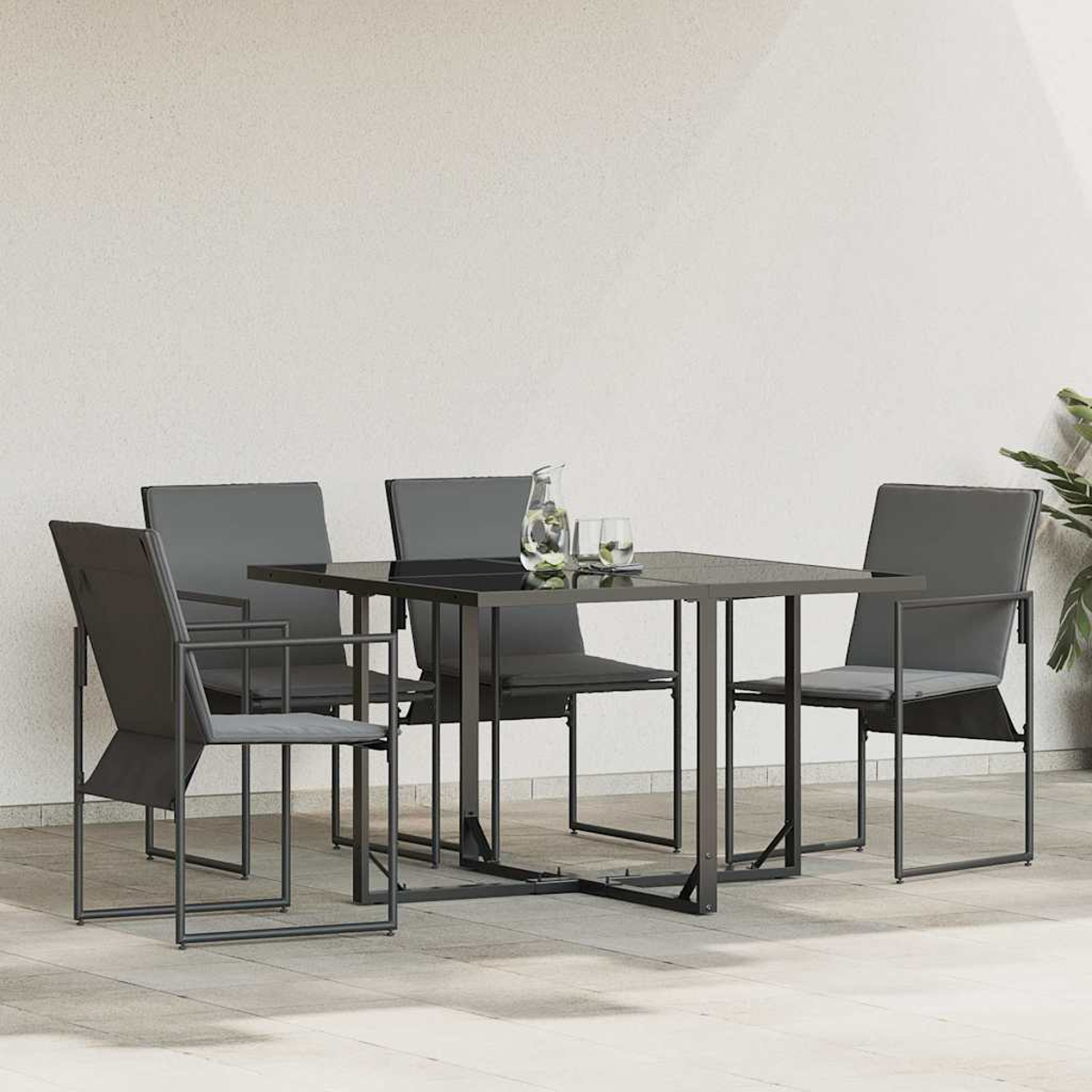 Ein quadratischer Tisch mit vier grauen Stühlen. Auf dem Tisch stehen ein Krug und Gläser. Rechts ist eine Pflanze. Die Wand ist weiß.