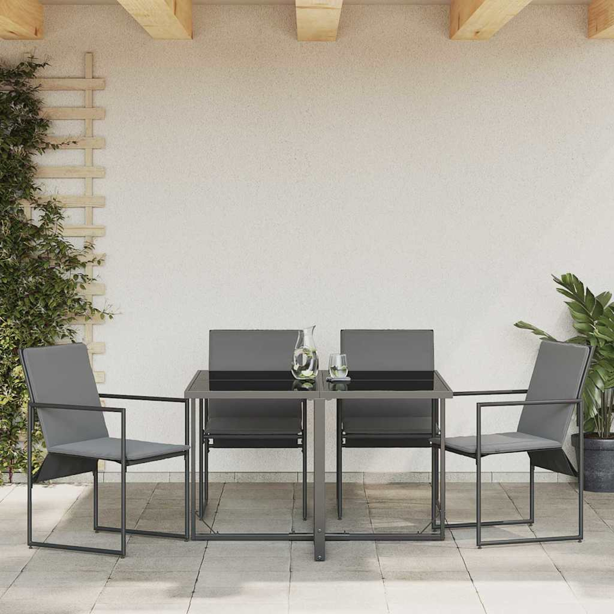 Essgruppe im Freien mit grauen Stühlen und einem schwarzen Tisch. Pflanzen an den Seiten und ein Holzbalken darüber. Eine Kanne und ein Glas auf dem Tisch.