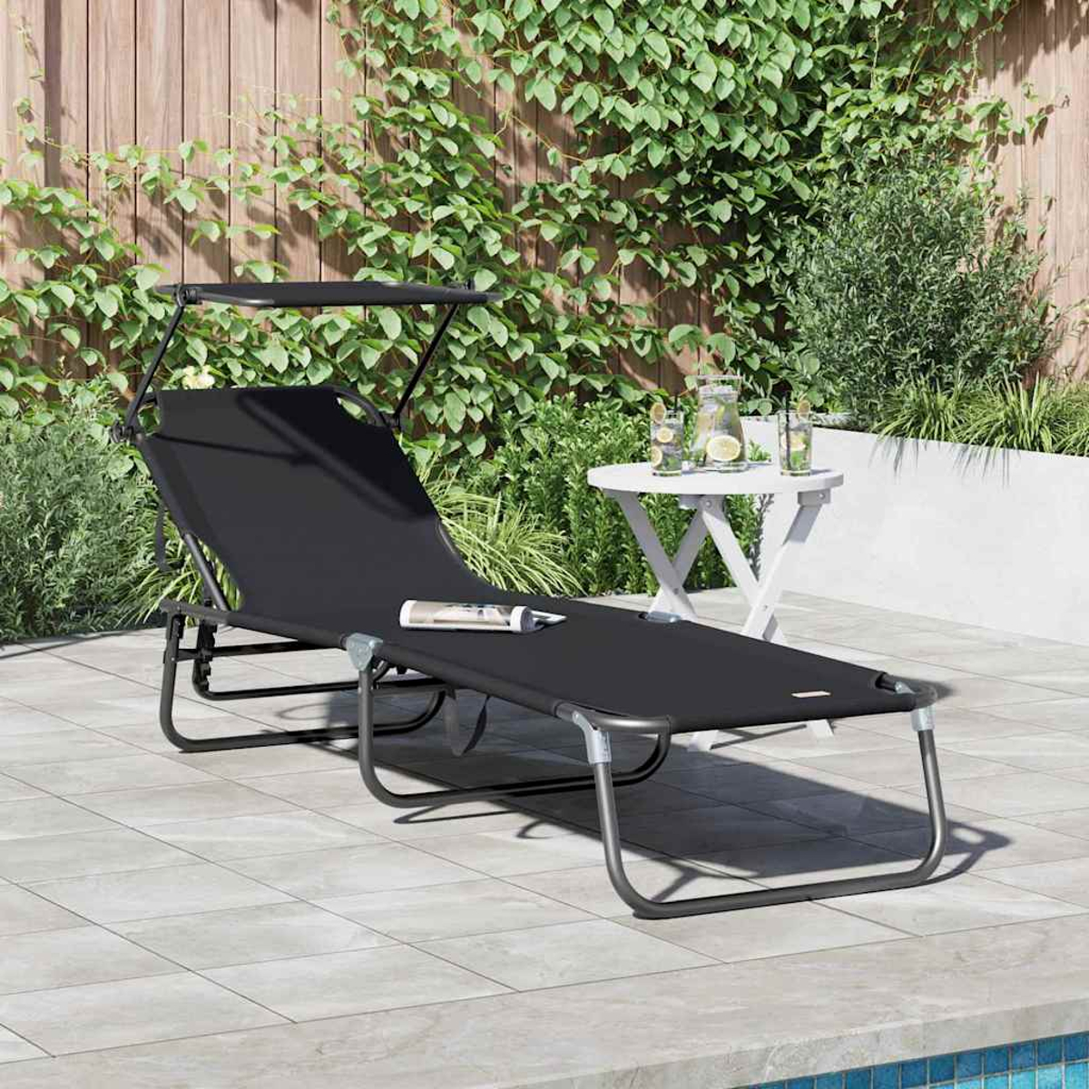 Eine schwarze Sonnenliege mit Baldachin steht auf einer Terrasse. Ein kleiner weißer Tisch mit Getränken ist in der Nähe, und ein Holzzaun ist im Hintergrund.