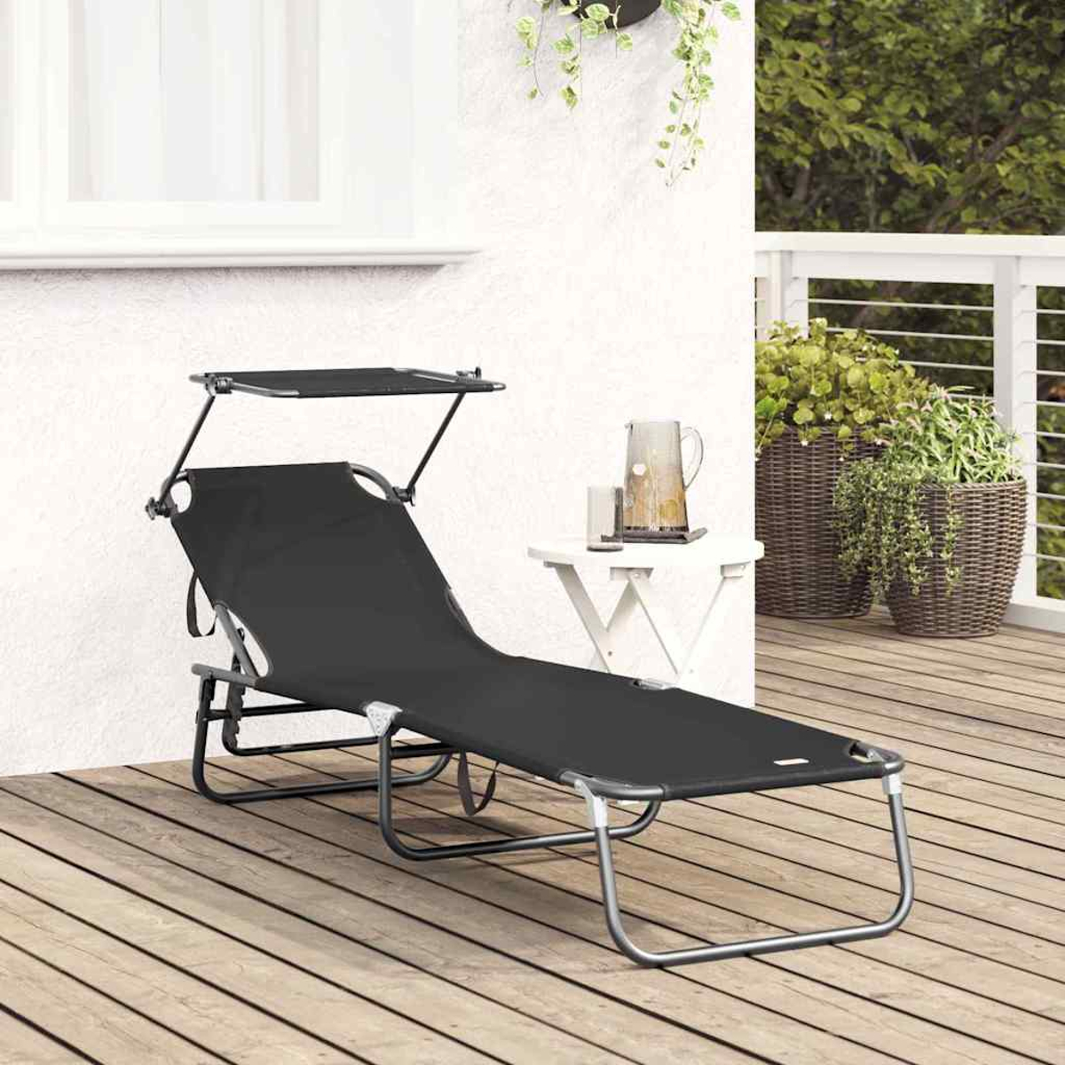 Eine schwarze Sonnenliege mit Sonnenschutz auf einer Holzterrasse. Ein weißer Tisch mit Kanne und Pflanzen steht daneben.
