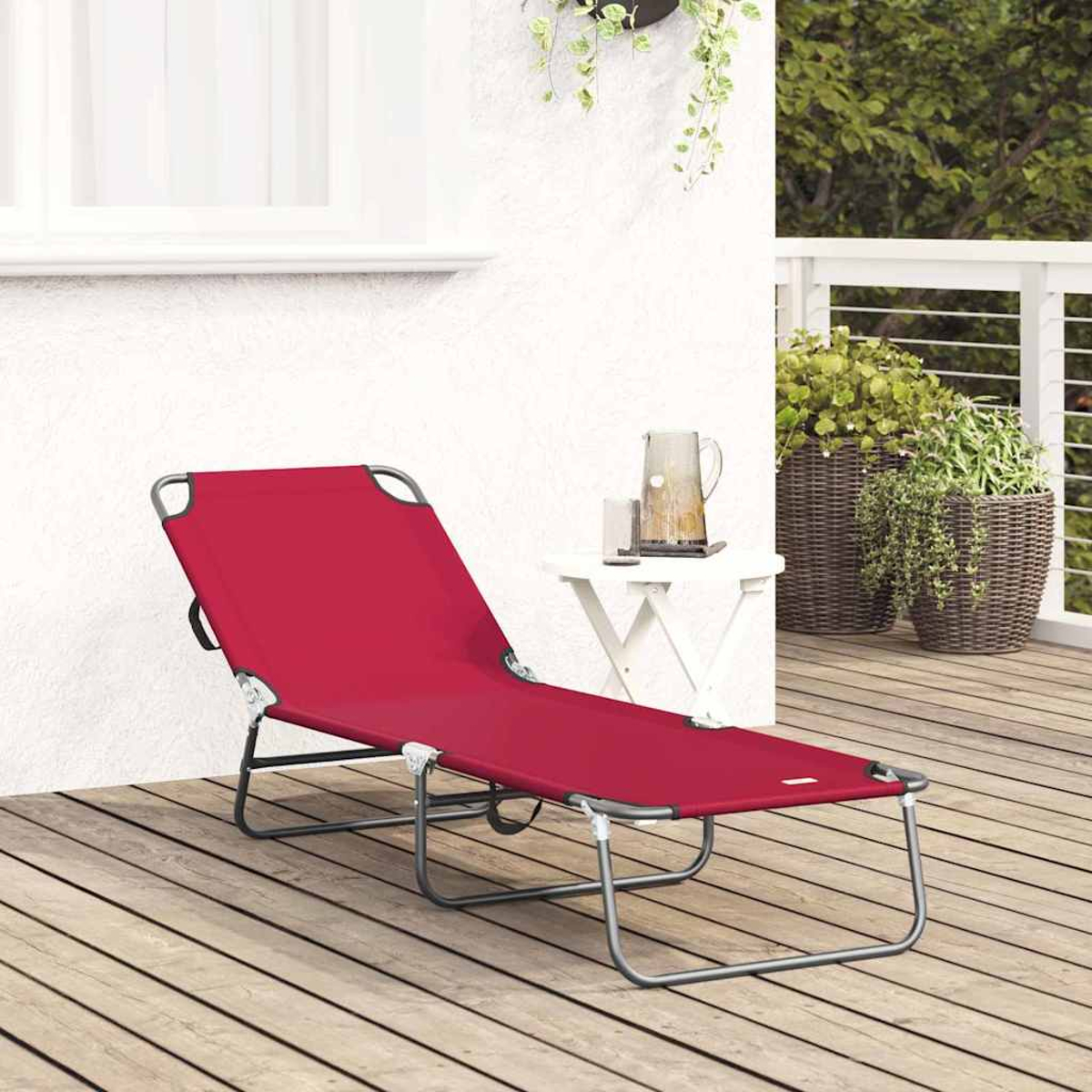 Ein roter Liegestuhl steht auf einer Holzterrasse. Ein kleiner weißer Tisch mit Kanne und Pflanzen steht rechts. Weiße Wand im Hintergrund.