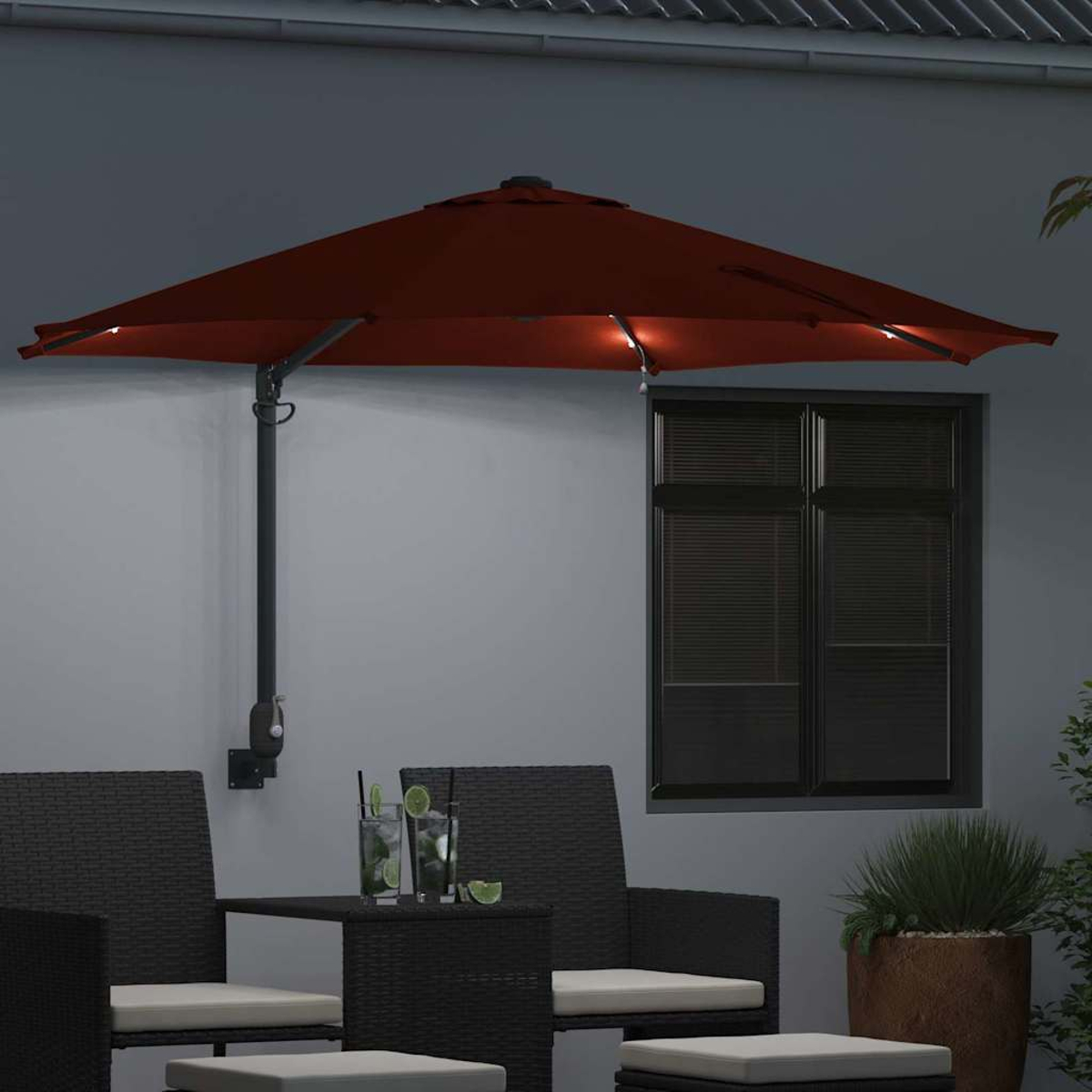 Un ombrellone rosso con luci è montato su un muro grigio. Sotto ci sono sedie, un tavolo e bevande. Una finestra è sullo sfondo.
