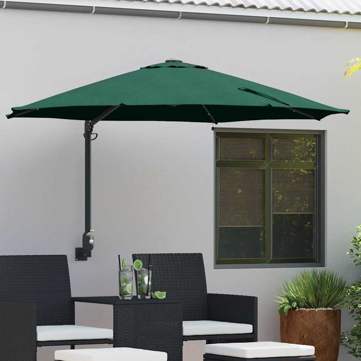 Un ombrellone verde da patio attaccato a un muro bianco ombreggia un tavolo e delle sedie. Le bevande sono sul tavolo. Una finestra è sullo sfondo.