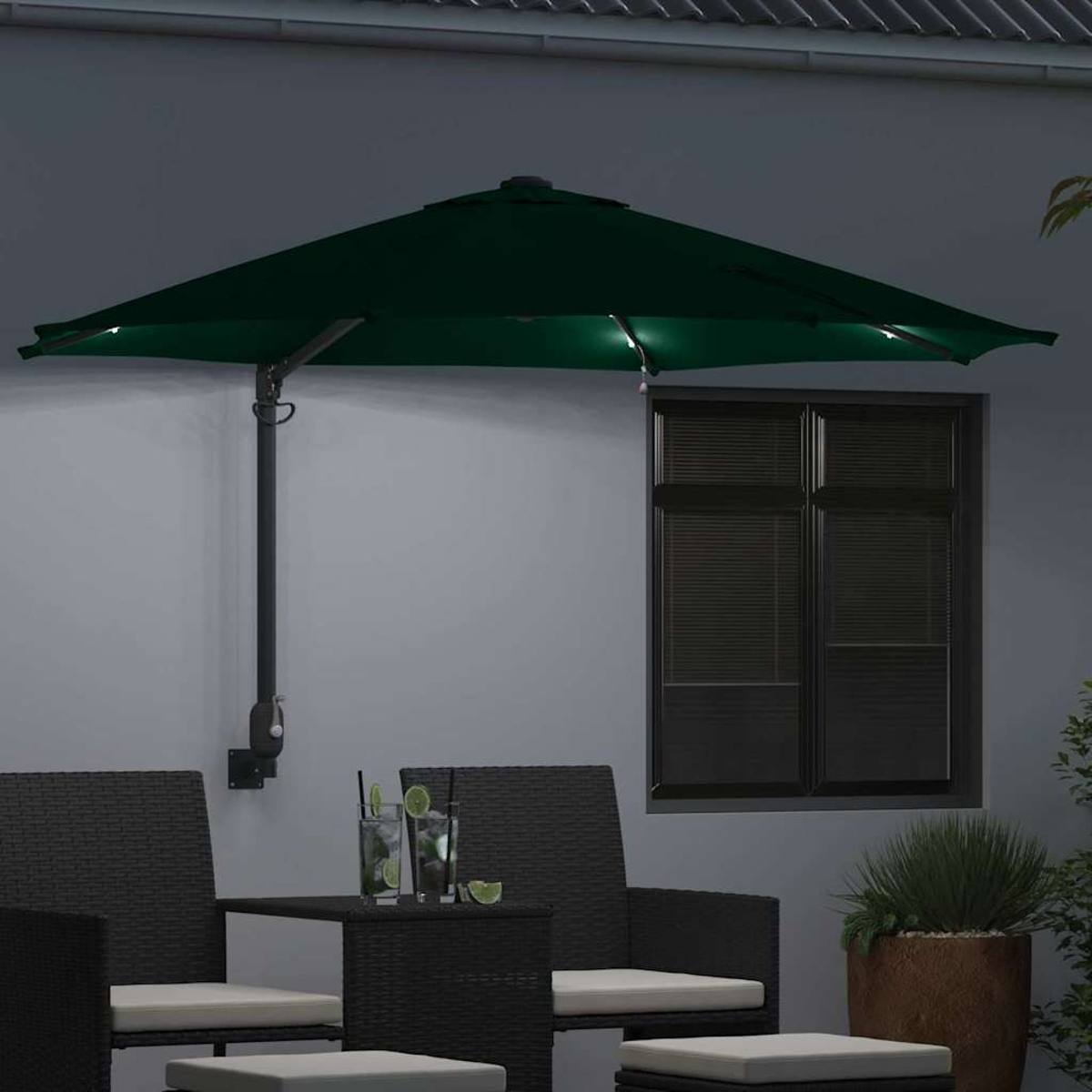Ombrellone da patio verde scuro con luci sopra mobili neri. Cocktail sul tavolo, finestra con persiane e pianta in vaso sullo sfondo.
