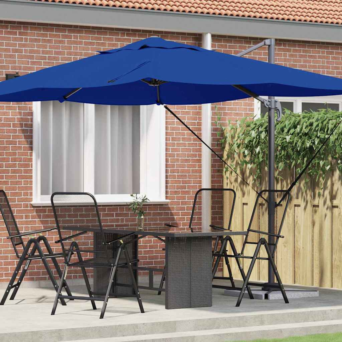 Arredamento patio con ombrellone blu, tavolo e sedie. Muro di mattoni sullo sfondo, recinzione in legno e foglie verdi.