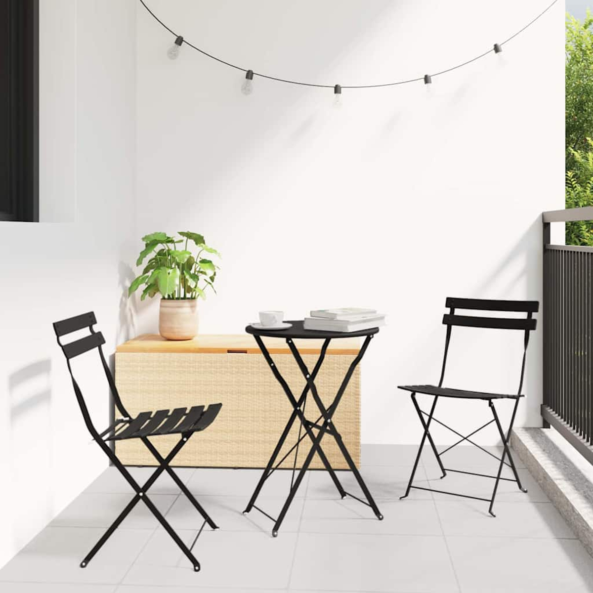 Eine Terrasse mit schwarzen Klappstühlen, einem kleinen Tisch und einer Pflanze. Lichterketten hängen darüber. Weiße Wände und Fliesenboden.