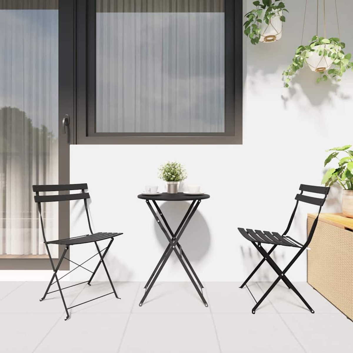 Schwarzes Bistro-Set auf einer Terrasse: Tisch und zwei Stühle. Fenster und Hängepflanzen im Hintergrund. Kleine Pflanze auf dem Tisch.