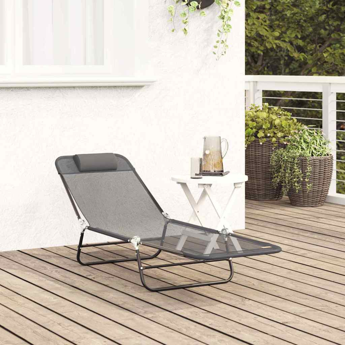 Ein grauer Liegestuhl steht auf einer Holzterrasse. Ein kleiner Tisch trägt eine Kanne und Gläser. Topfpflanzen sind in der Nähe.