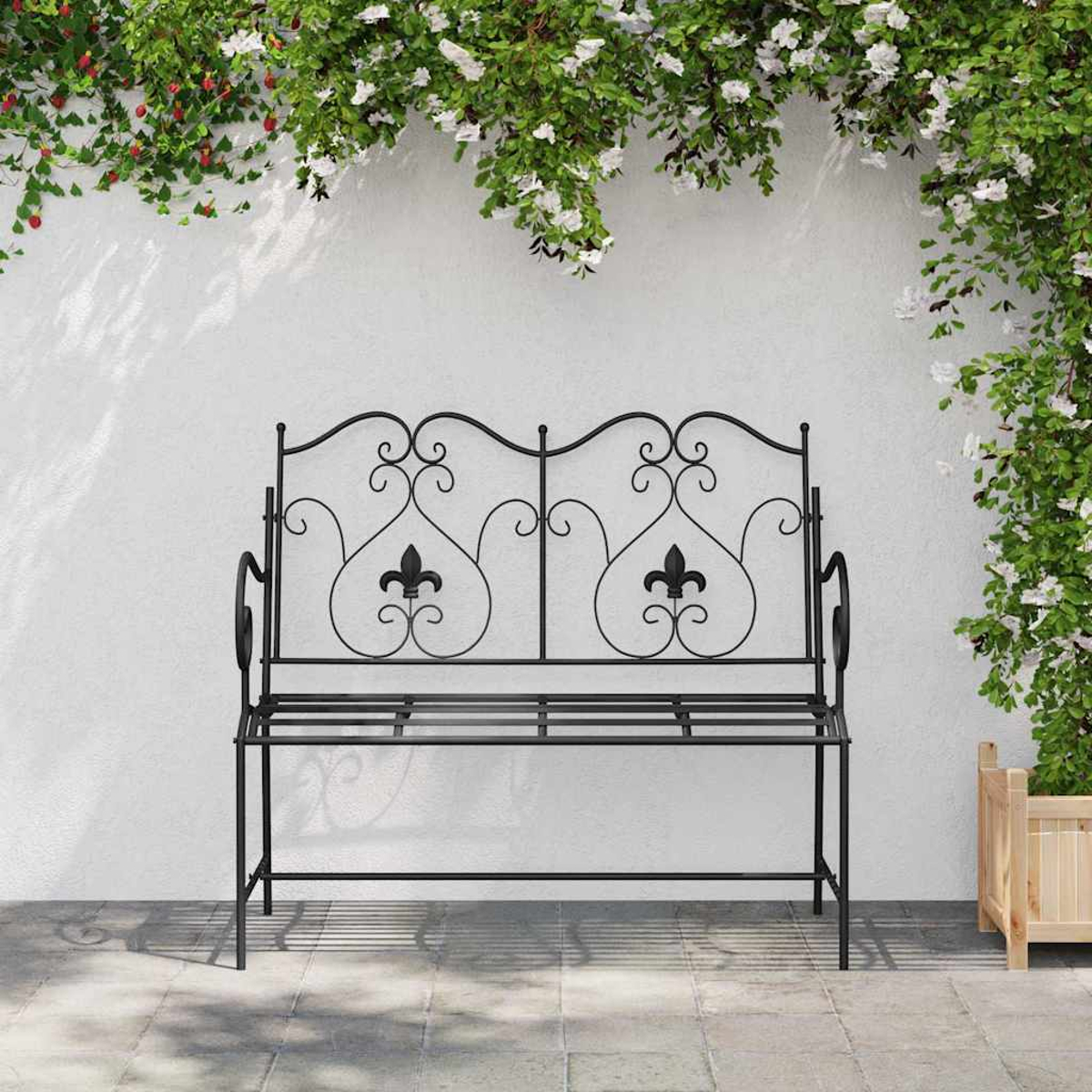 Banco negro de hierro forjado con diseño de flor de lis, contra una pared blanca con plantas trepadoras y flores.