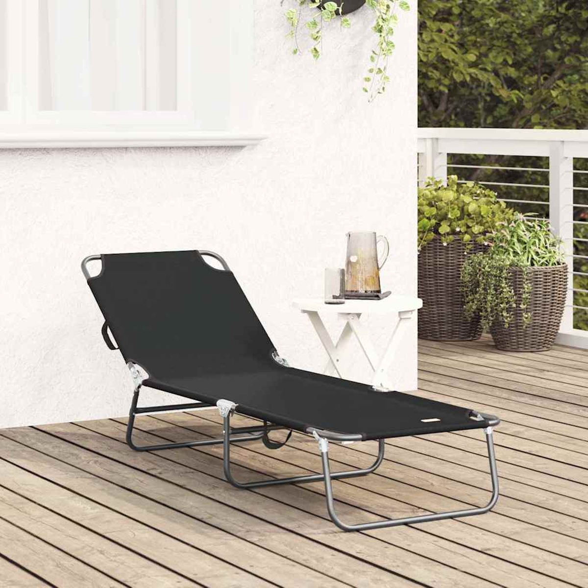 Eine schwarze Liege auf einer Holzterrasse. Ein kleiner weißer Tisch trägt einen Krug. Grüne Pflanzen im Hintergrund.