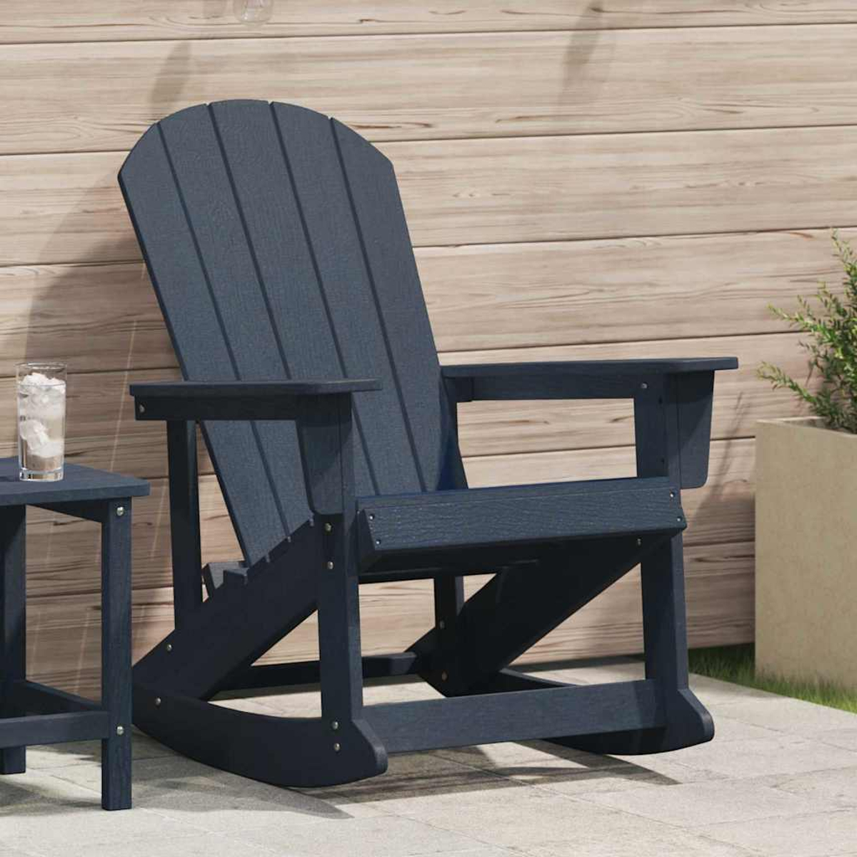 Ein marineblauer Schaukelstuhl auf einer Terrasse. Ein kleiner Tisch mit einem Glas Wasser steht daneben. Holzwand im Hintergrund.
