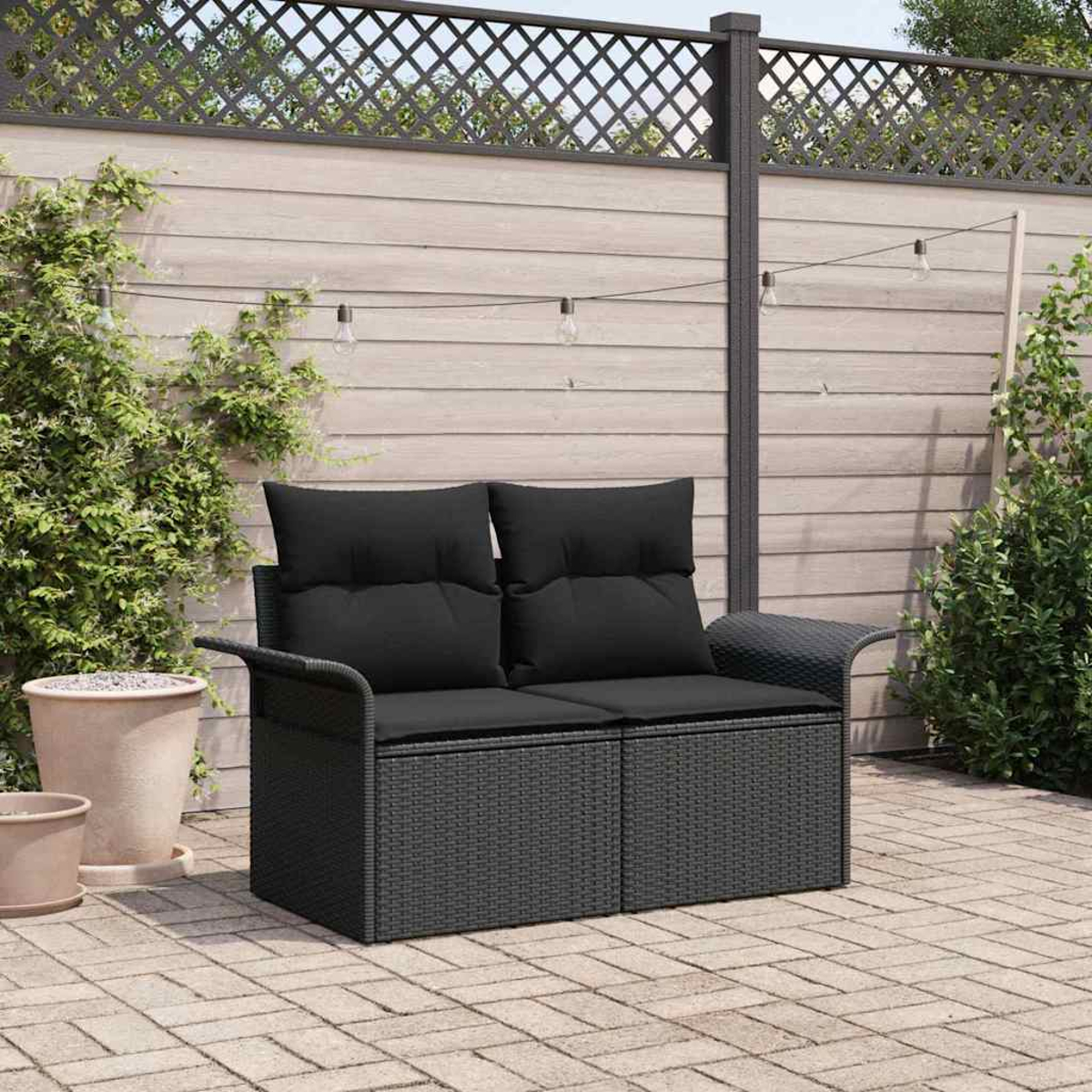 Ein schwarzes Outdoor-Sofa aus Korbgeflecht mit schwarzen Kissen steht auf einer Ziegelterrasse. Grüne Pflanzen und ein Holzzaun sind im Hintergrund.