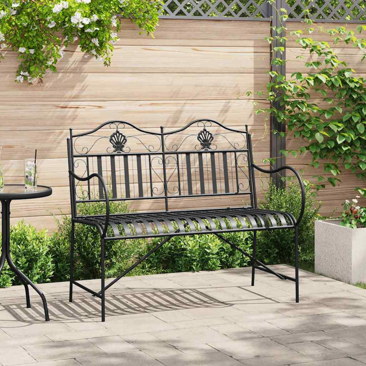 Schwarze Metallbank auf einer Terrasse. Dahinter ein Holzzaun mit Kletterpflanzen. Links ein kleiner Tisch.