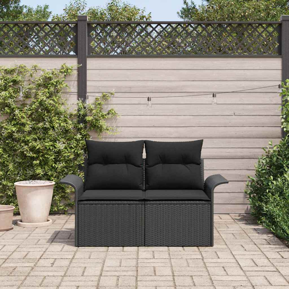 Schwarzes Korbmöbel mit Kissen auf einer Ziegelterrasse, mit Holzzaun im Hintergrund und grünen Pflanzen.