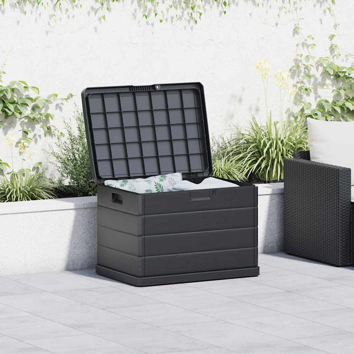 Offene schwarze Truhe auf einer Terrasse. Innen Kissen. Pflanzen und ein Stuhl sind im Hintergrund. Weiße Wand.