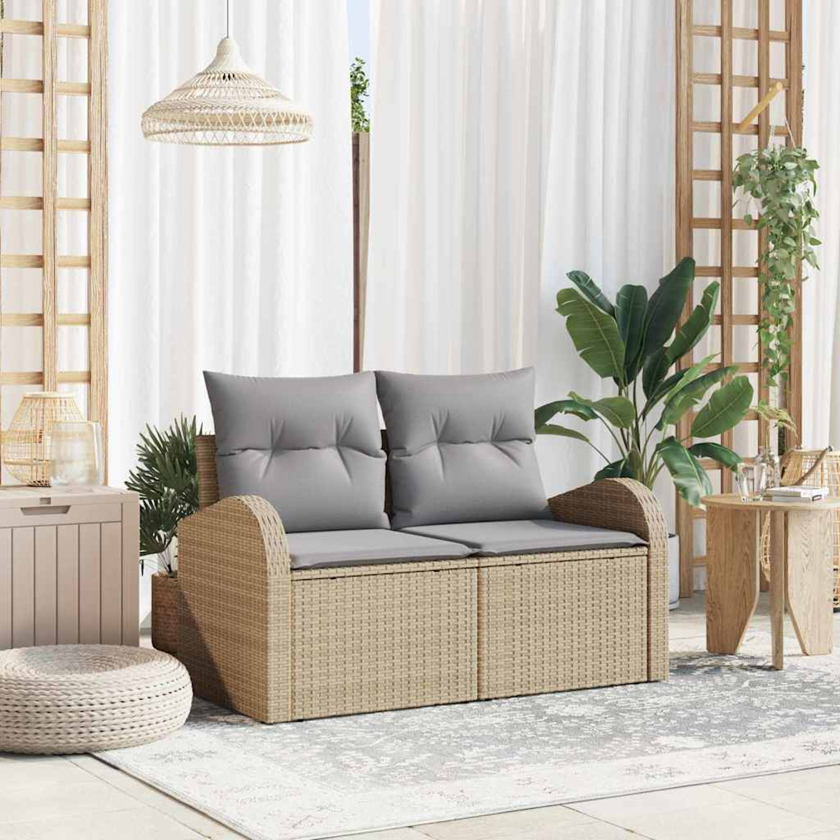 Ein Korbmöbel mit grauen Kissen steht auf einem gemusterten Teppich. Pflanzen und Holzmöbel umgeben das Sofa, im Hintergrund weiße Vorhänge.