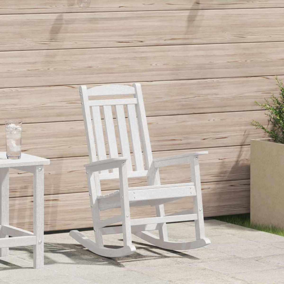 Ein weißer Schaukelstuhl steht auf einer Terrasse neben einem kleinen Tisch mit einem Glas Wasser. Holzwand im Hintergrund.