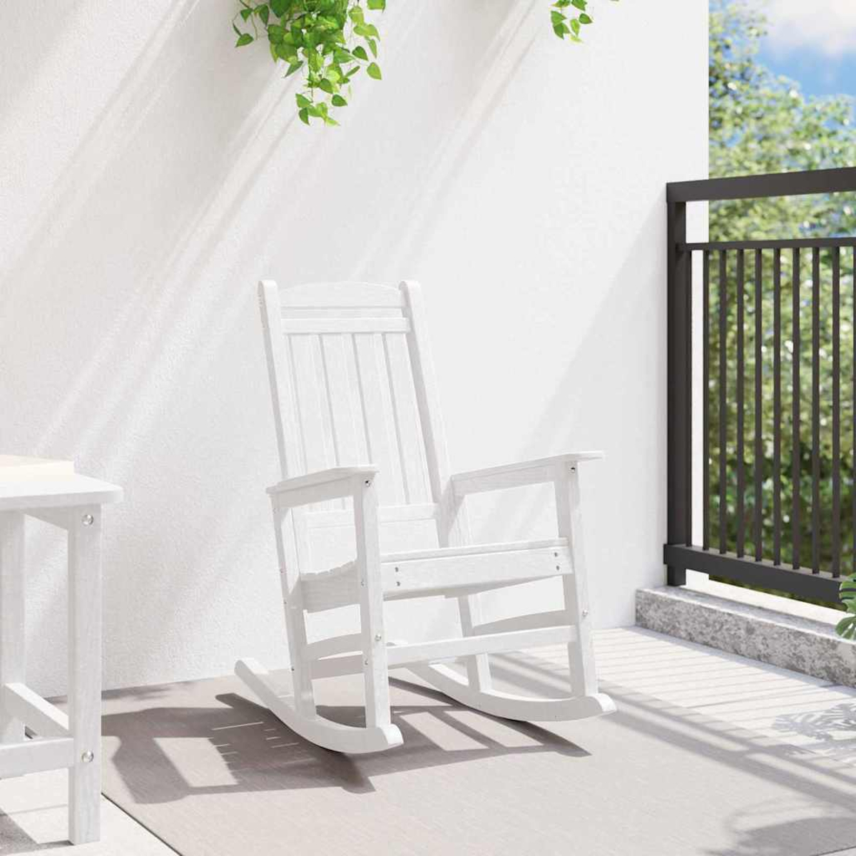 Ein weißer Schaukelstuhl auf einer Terrasse. Grüne Pflanzen hängen darüber. Links steht ein Tisch. Rechts befindet sich ein schwarzes Geländer.