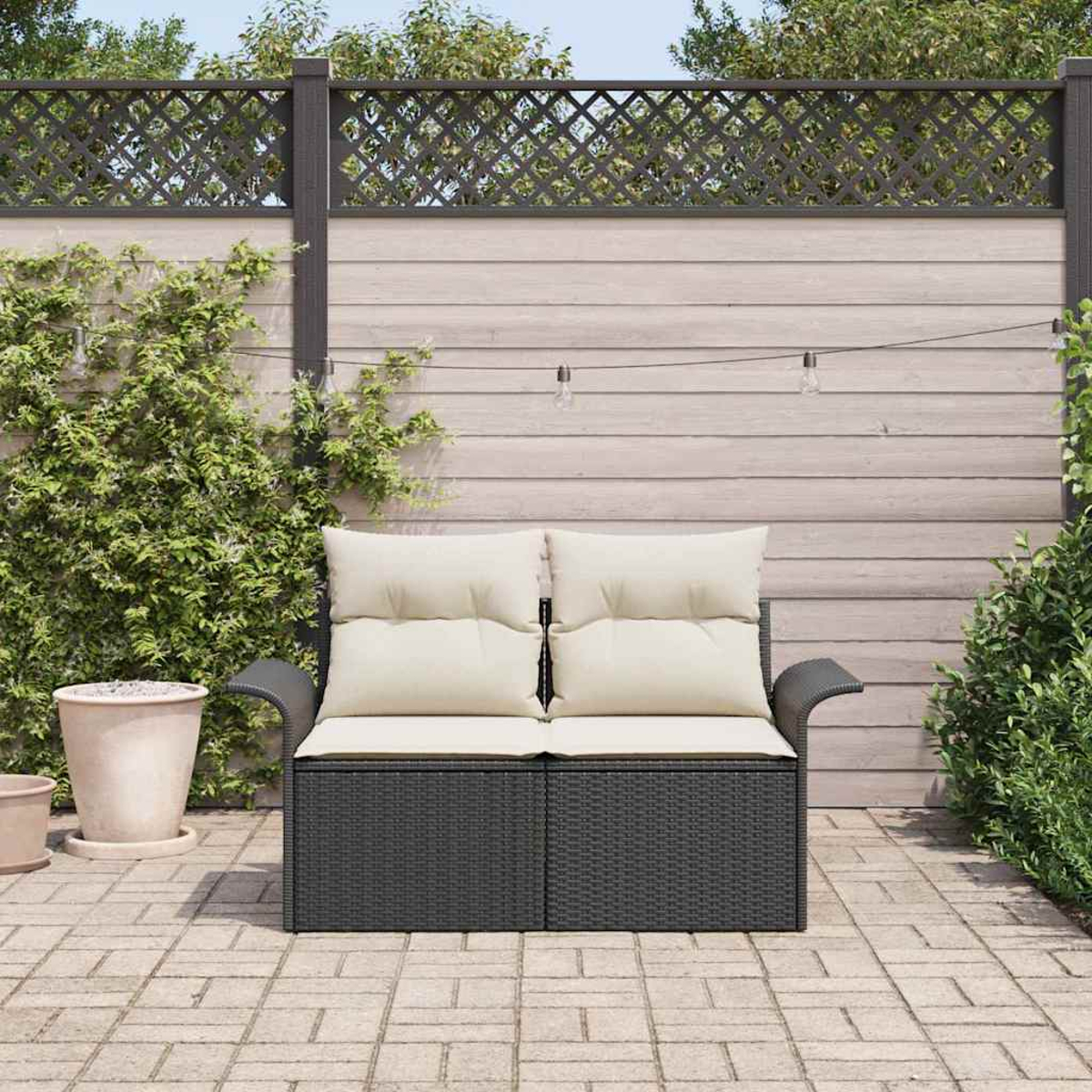 Ein dunkelgraues Korbmöbel mit cremefarbenen Kissen steht auf einer gepflasterten Terrasse. Dahinter ein Holzzaun mit Kletterpflanzen.