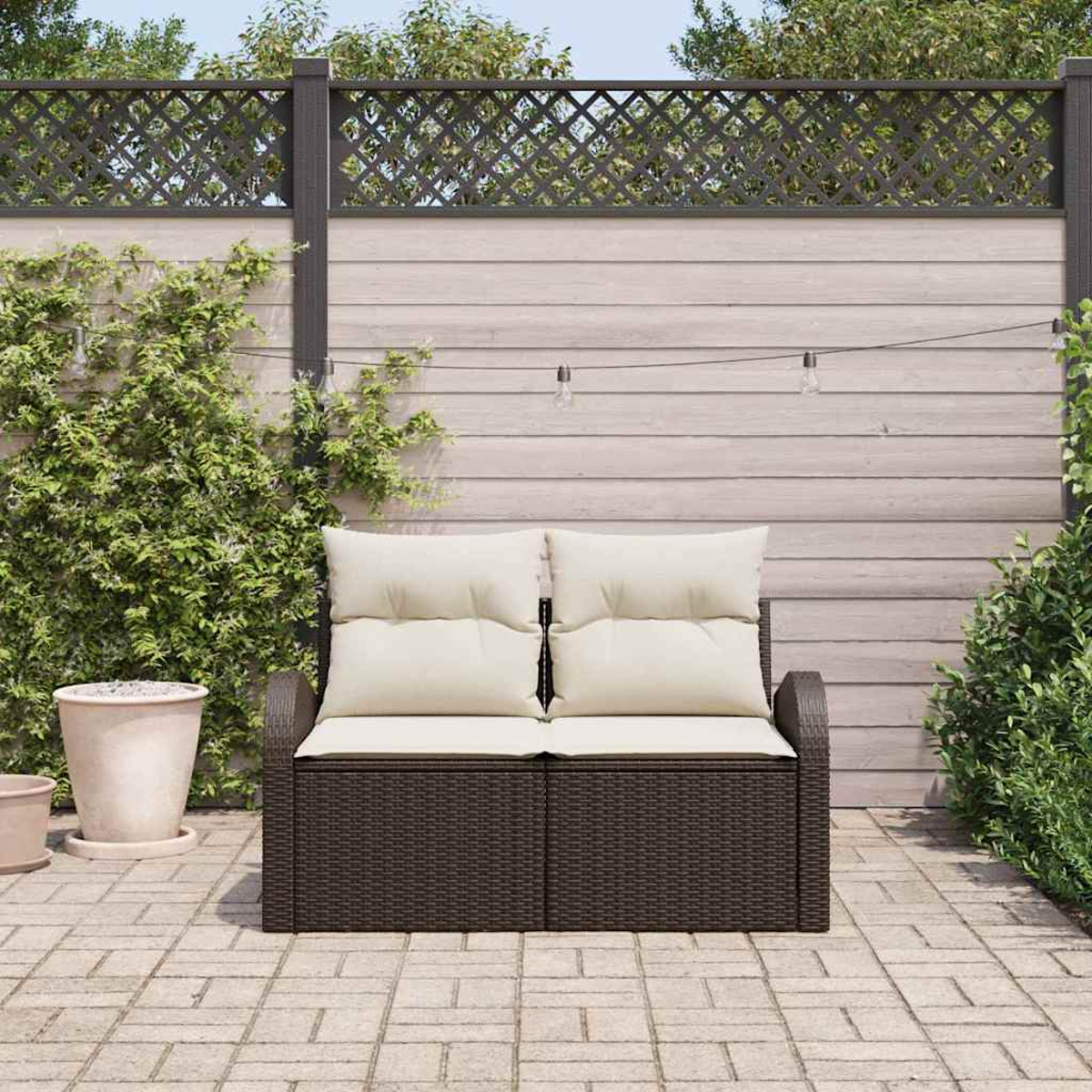 Ein braunes Rattan-Sofa mit cremefarbenen Kissen auf einer Ziegelterrasse, mit Holzzaun und Grün im Hintergrund.