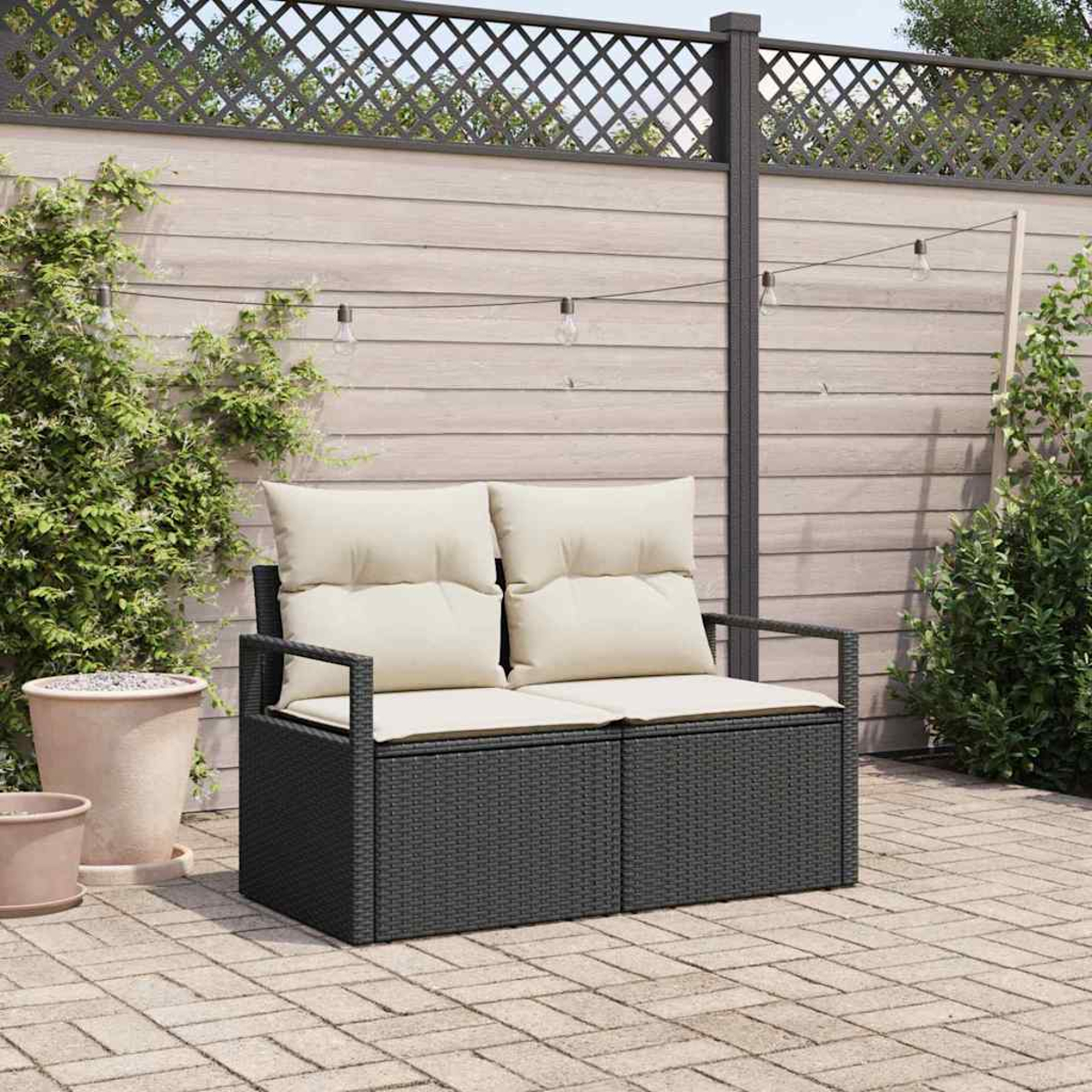 Ein schwarzes Korbmöbel mit cremefarbenen Kissen steht auf einer Backstein-Terrasse. Dahinter ein Holzzaun mit Kletterpflanzen und dekorativen Lichtern.
