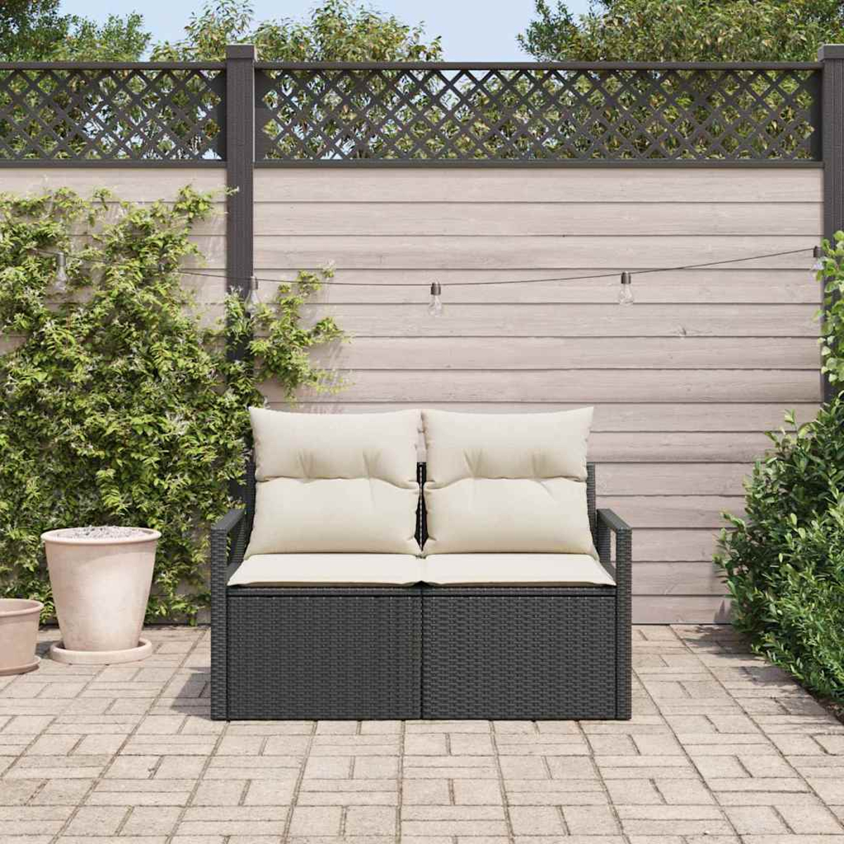 Ein dunkelgraues Outdoor-Liebesofa mit cremefarbenen Kissen steht auf einer Ziegelsteinterrasse. Grüne Pflanzen und ein Holzzaun sind im Hintergrund.