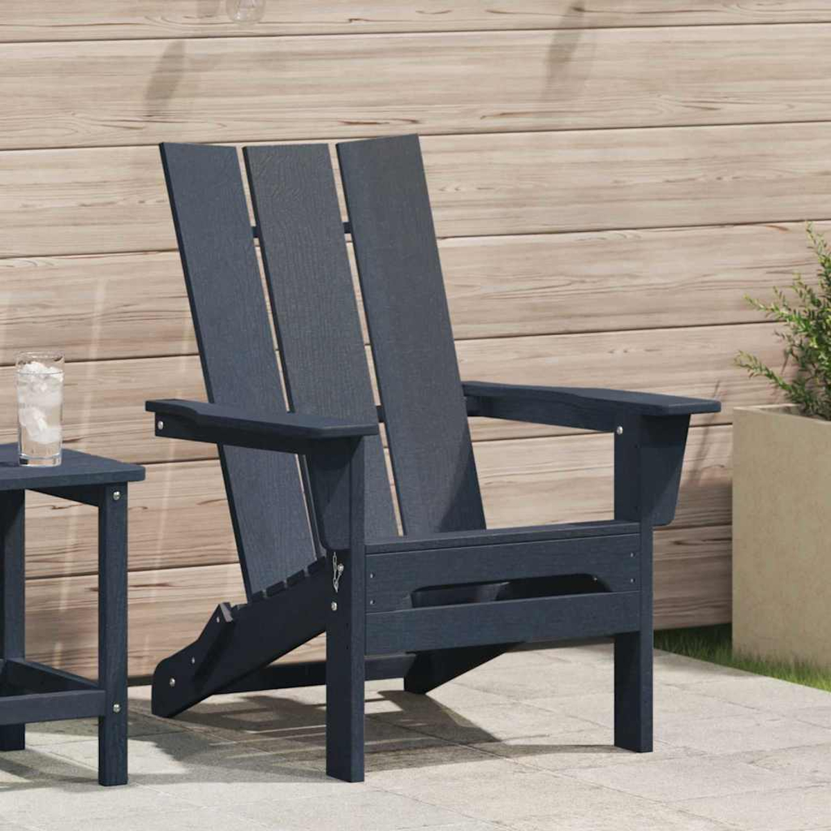 Una silla Adirondack azul marino está en un patio. Una mesa auxiliar con un vaso de agua con hielo está al lado. Una pared de madera en el fondo.