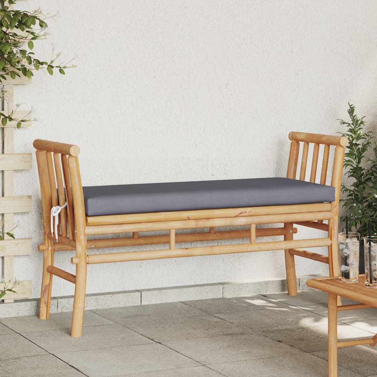 Eine Bambusbank mit grauem Kissen steht auf einer Terrasse. Rechts steht ein kleiner Tisch mit zwei Gläsern. Im Hintergrund Grün.
