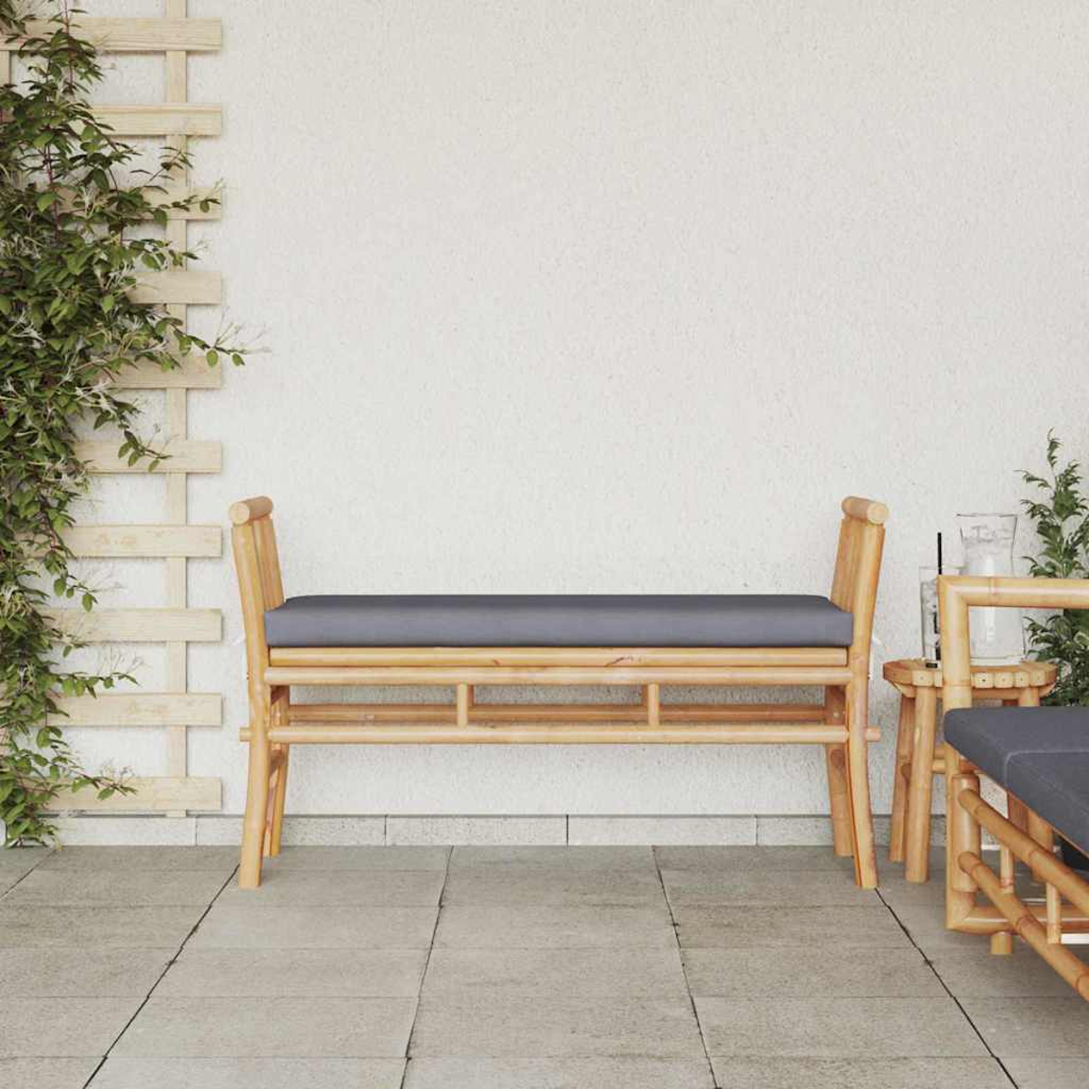 Eine Bambusbank mit grauem Kissen steht auf einer gefliesten Terrasse. Links ein Spalier mit grünen Weinreben. Rechts ein kleiner Tisch und Stuhl.