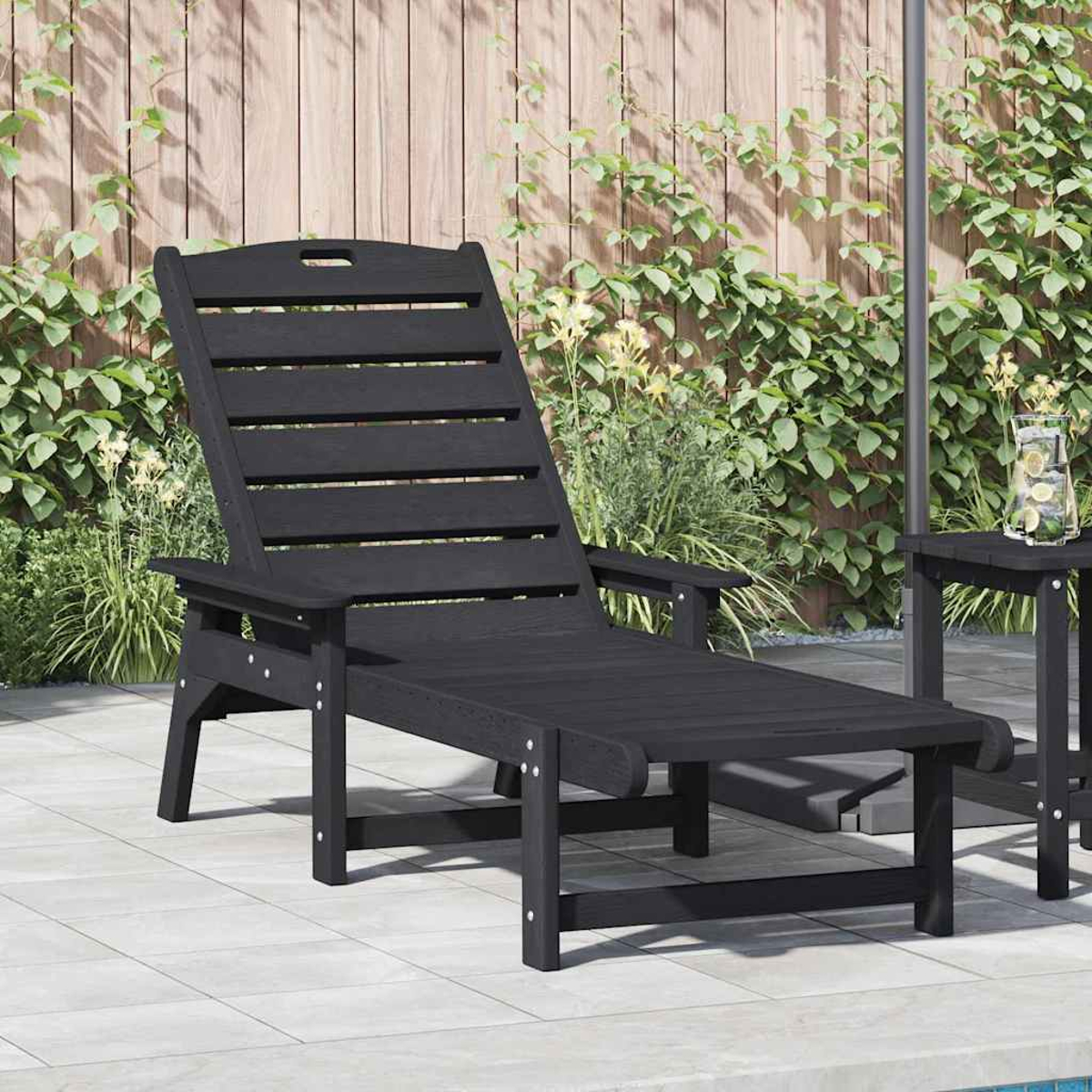 Una tumbona de madera negra en un patio. Al fondo, una valla de madera cubierta de enredaderas. A la derecha, una mesa pequeña.