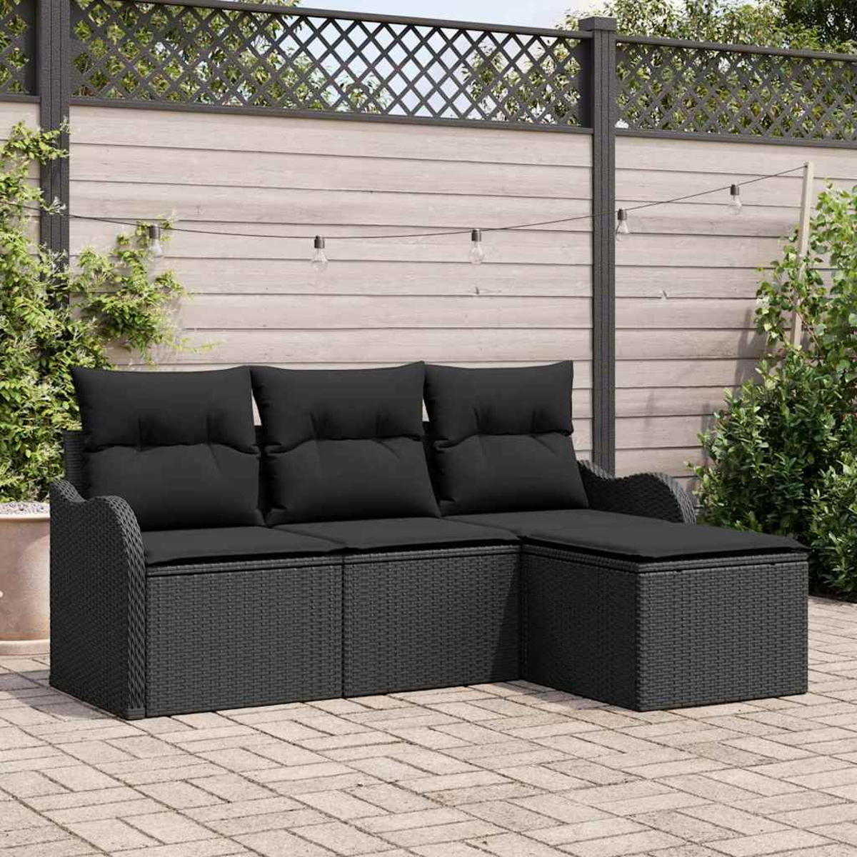 Schwarzes Rattan-Ecksofa mit Kissen auf einer Terrasse. Im Hintergrund befinden sich ein Holzzaun und Lichterketten. Pflanzen sind an den Seiten.