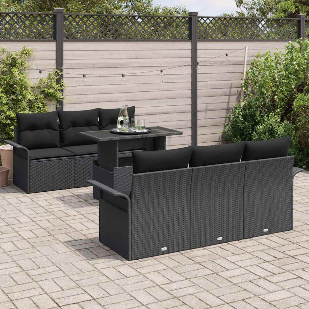 Szene im Freien mit dunklen Korbmöbeln: Sofa, Sessel und Tisch. Schwarze Kissen auf den Sitzen. Ein Zaun und Grün im Hintergrund.