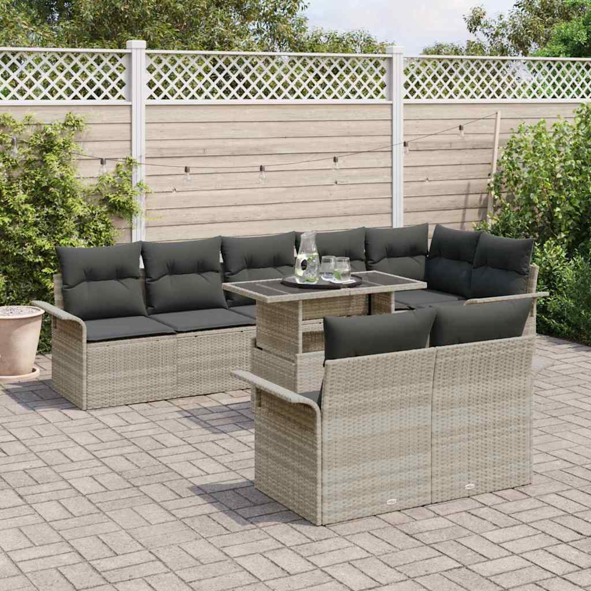 Garten-Terrassen-Set: graue Kissen auf Rattanmöbeln, Tisch mit Getränken, Ziegel-Terrasse, weißer Zaun, Grün.