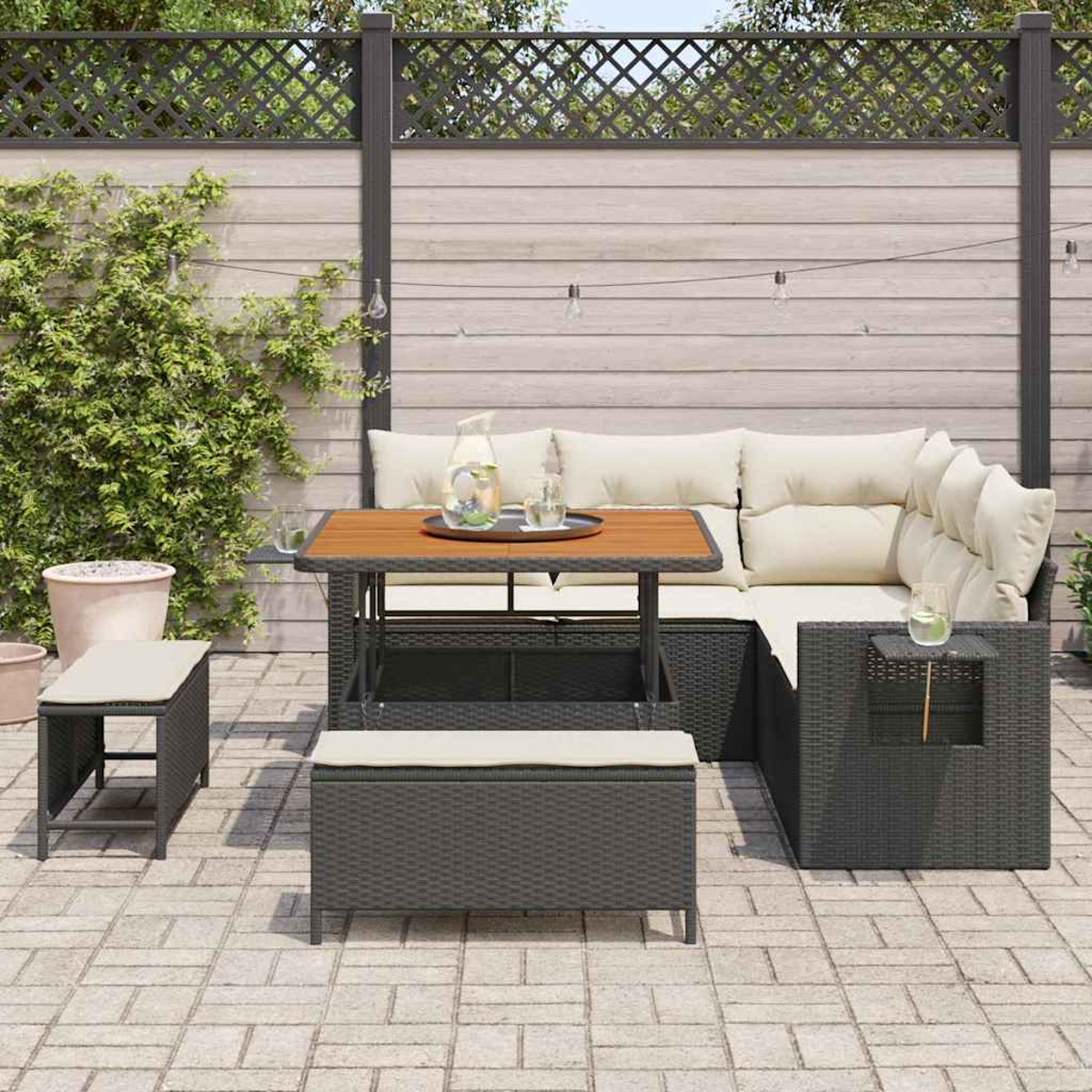 Set da patio esterno con tavolo, panche e divano componibile. Una caraffa d'acqua, bicchieri e cuscini adornano il set. Lo sfondo mostra una recinzione.