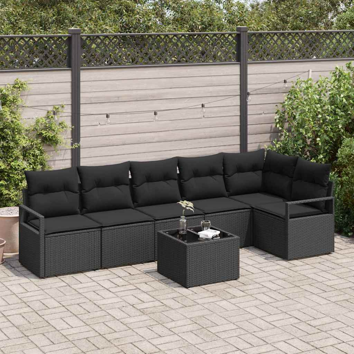 Schwarzes Outdoor-Ecksofa aus Korbgeflecht mit schwarzen Kissen, quadratischem Tisch und Pflanzen auf einer Backsteinterrasse mit Zaun.