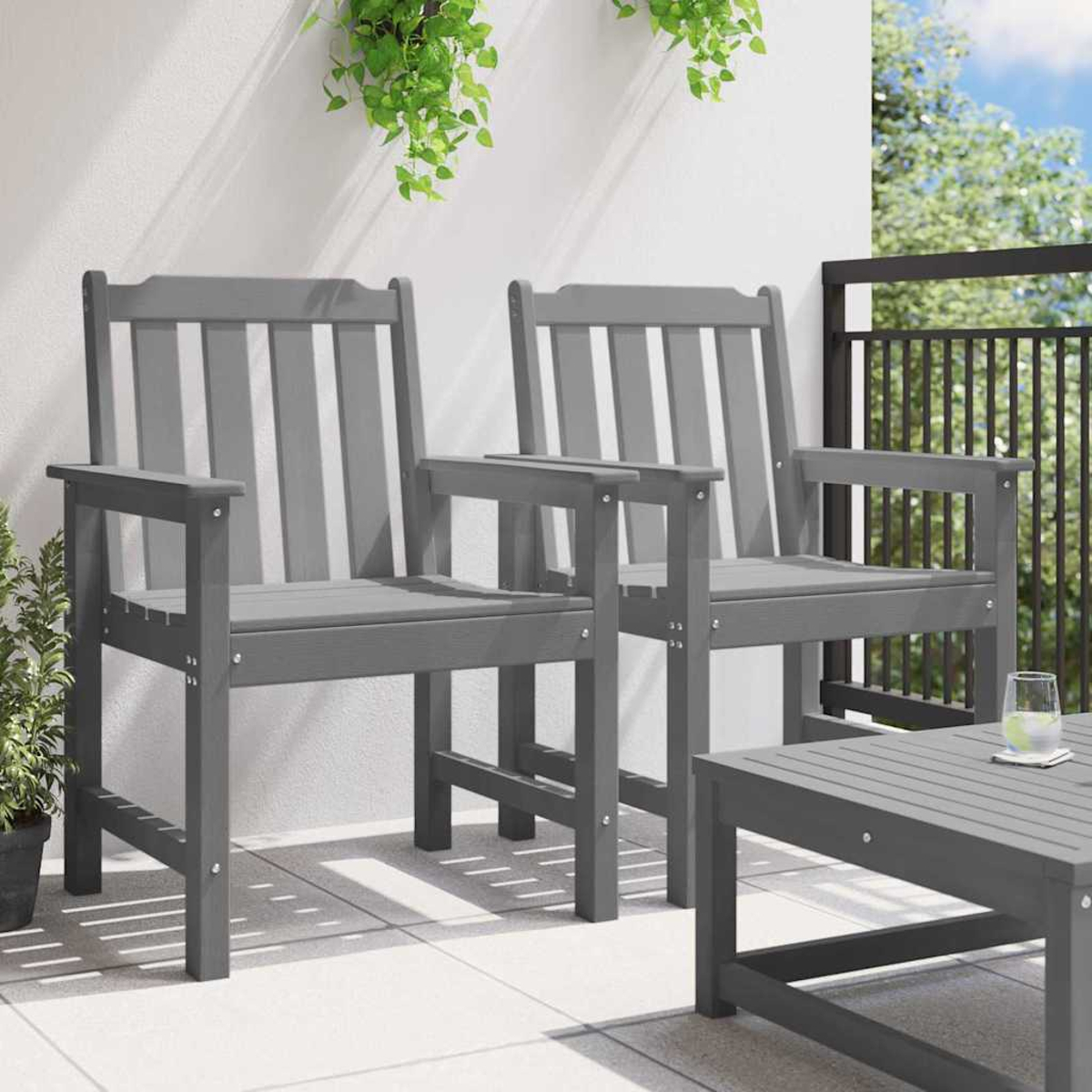 Zwei graue Holzstühle auf einer Terrasse. Ein kleiner Tisch und ein Glas mit Wasser. Grüne Pflanzen im Hintergrund und oben.