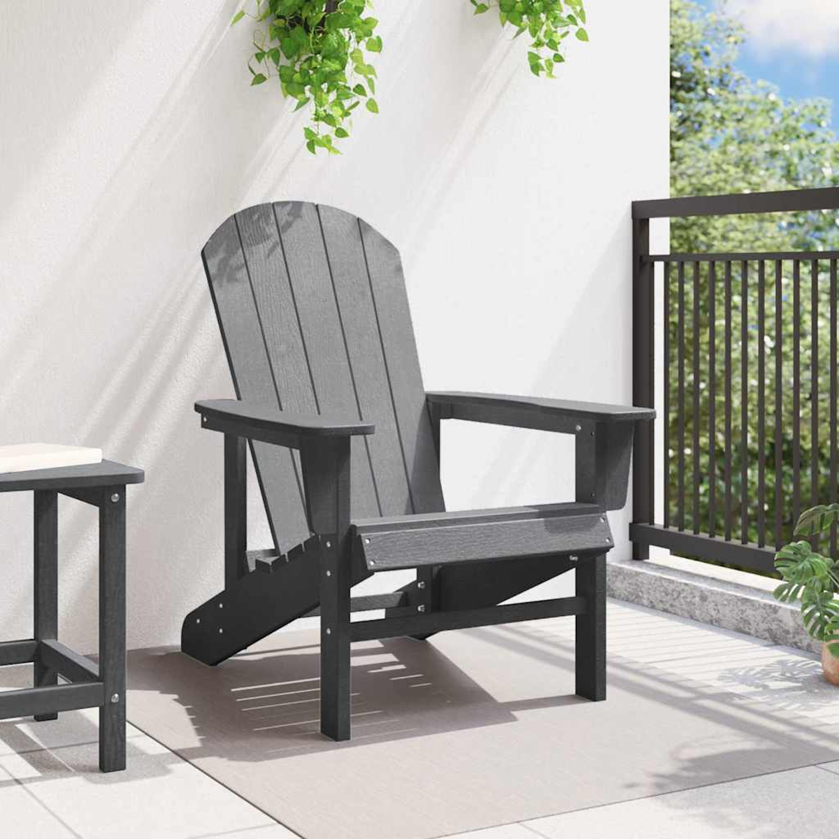 Ein grauer Adirondack-Stuhl auf einer Terrasse. Links steht ein kleiner Tisch. Grüne Pflanzen hängen darüber. Im Hintergrund ist ein schwarzes Geländer.