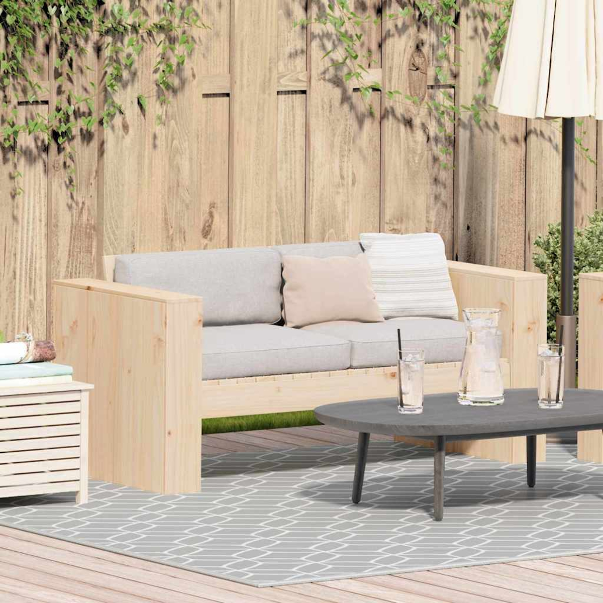 Escena al aire libre con sofá de madera, mesa y alfombra. Hiedra en valla de madera. Vasos y jarra de agua en la mesa.