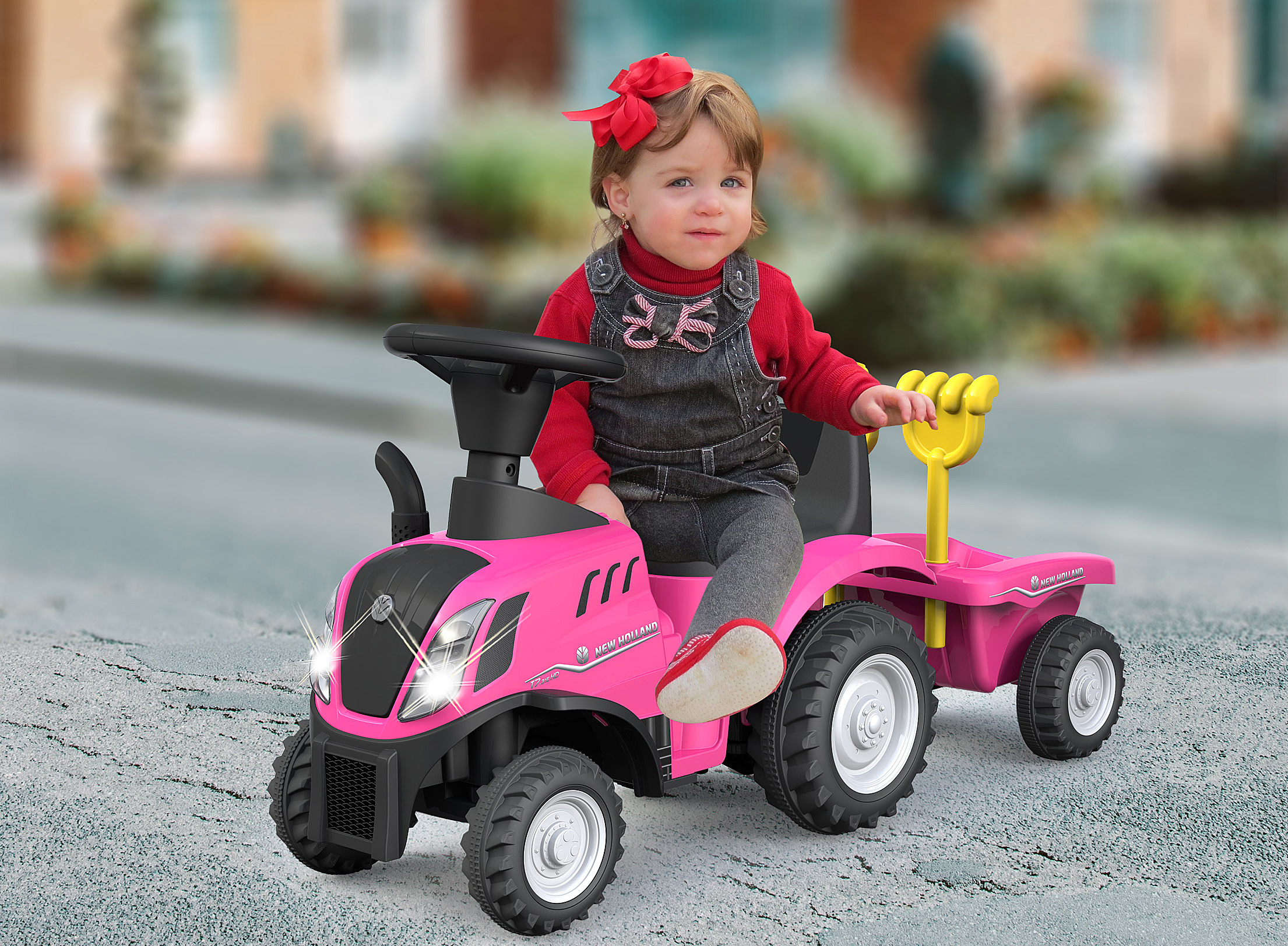 Un niño pequeño está sentado en un tractor de juguete rosa, con un lazo rojo, una camisa roja y un mono. El tractor tiene un remolque.