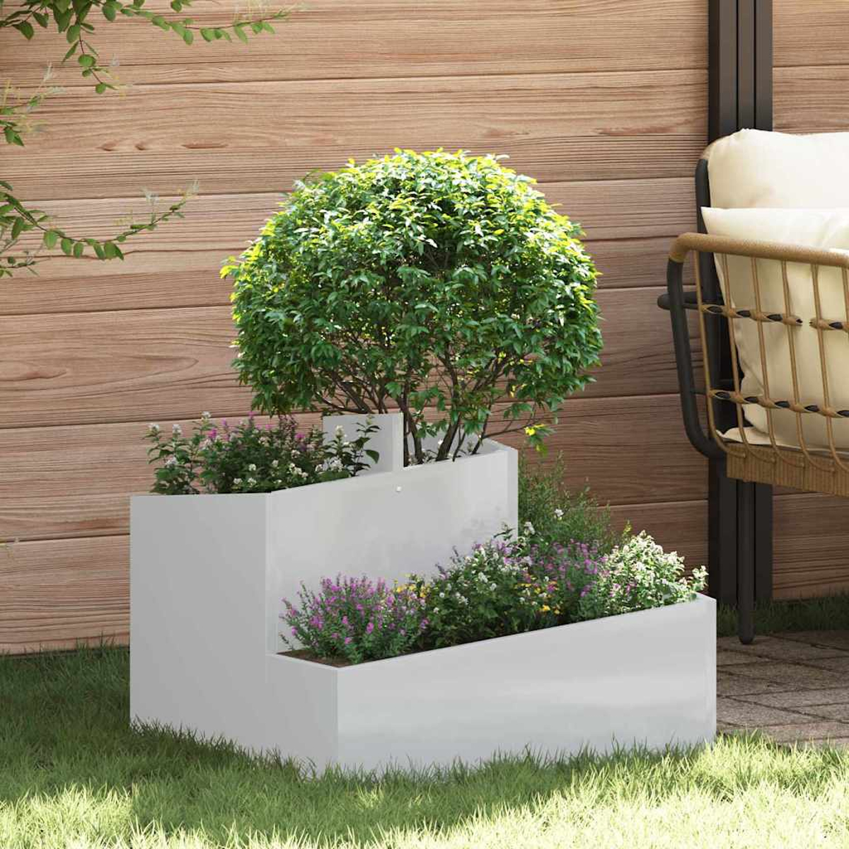 Una fioriera bianca a più livelli con piante verdi e fiori è appoggiata sull'erba. Una staccionata di legno e una sedia con cuscini sono sullo sfondo.