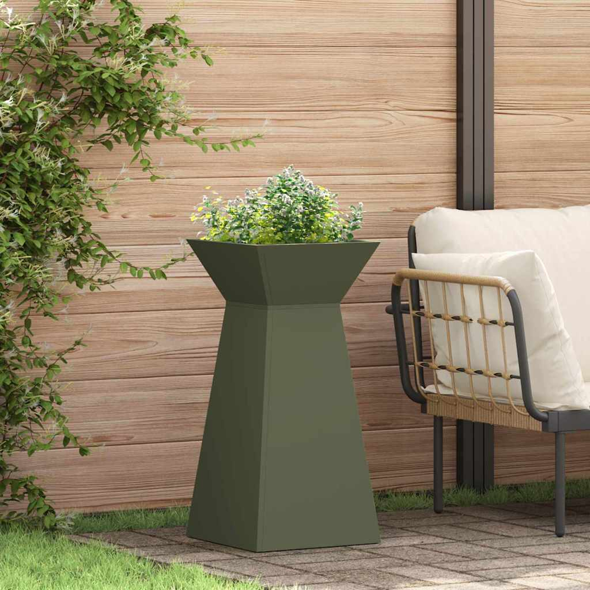 Una fioriera verde oliva con piante è su un patio in mattoni. Una staccionata in legno e una sedia con un cuscino sono sullo sfondo.