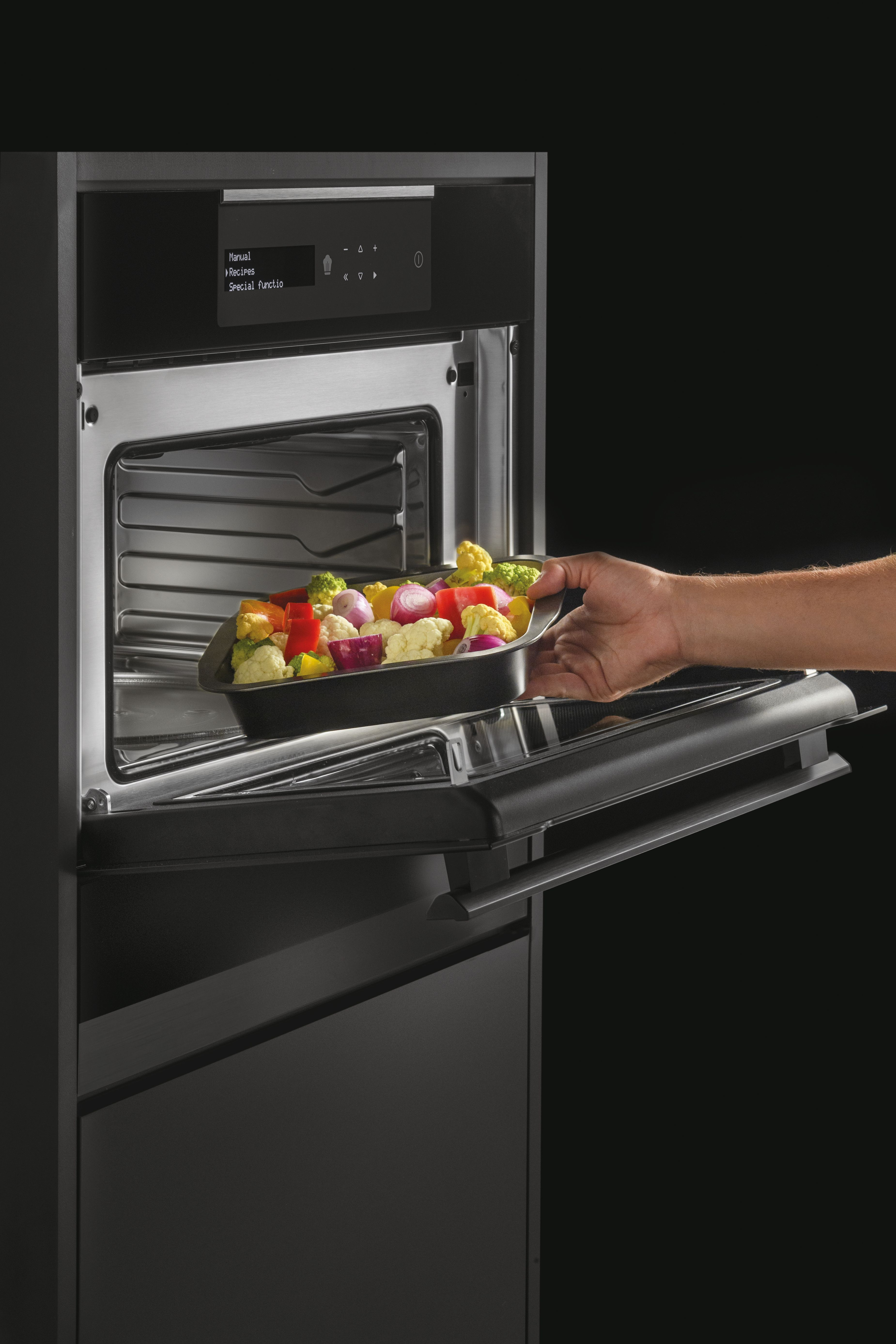 Mano colocando bandeja de vegetales coloridos en horno moderno. Pantalla muestra ajustes. Fondo negro.