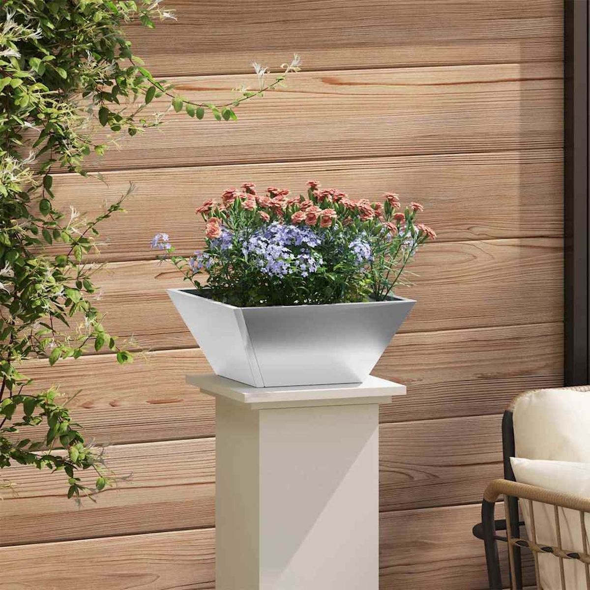 Una fioriera quadrata in metallo con fiori su un piedistallo bianco. Parete in legno dietro, viti verdi a sinistra e una sedia a destra.