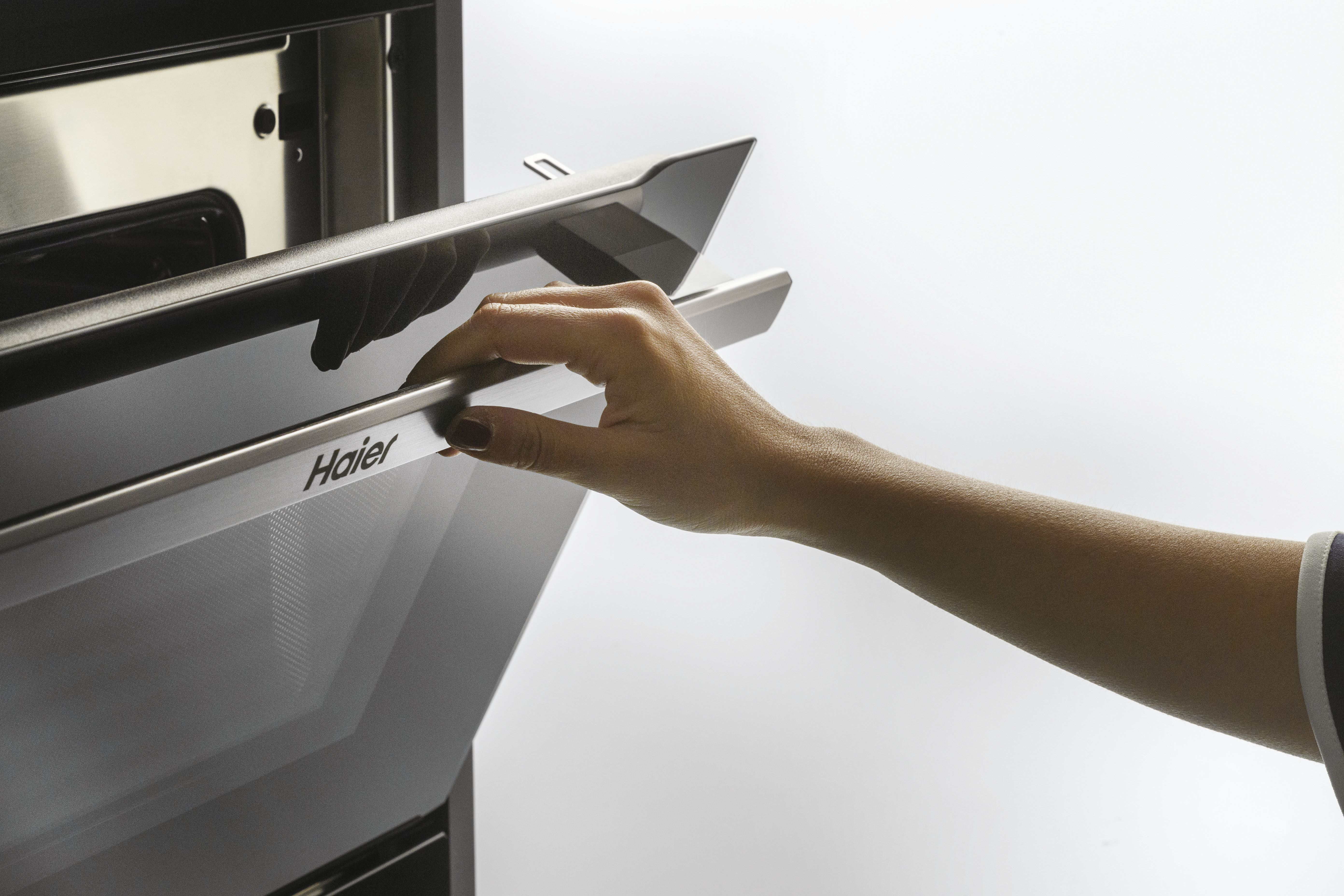 Una mano abre un horno. El horno es negro y plateado, con la marca 'Haier' visible.