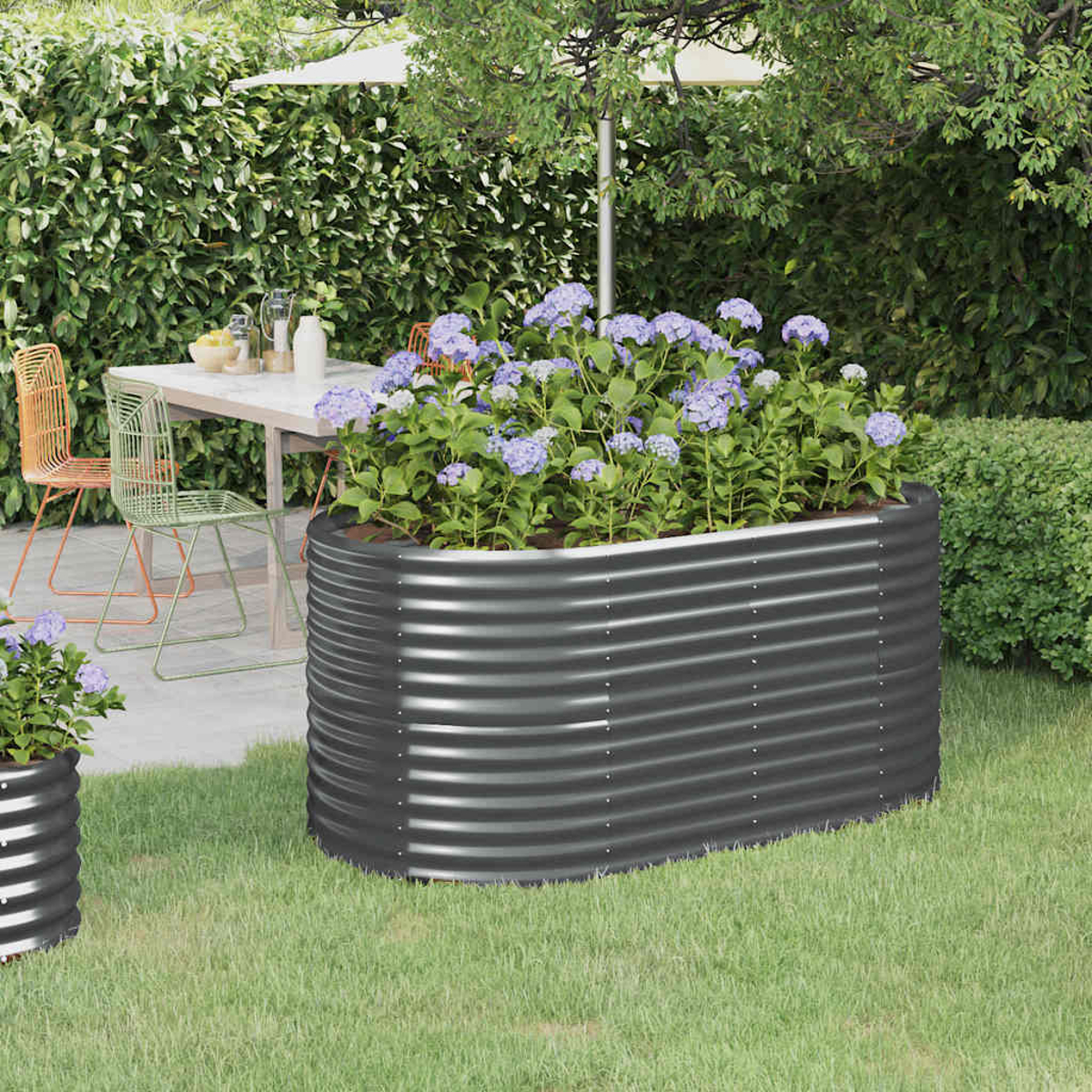 Ein Metallpflanzer mit lila Blumen steht auf Gras. Dahinter ein Tisch mit Stühlen und eine Hecke. Ein Schirm beschattet die Szene.