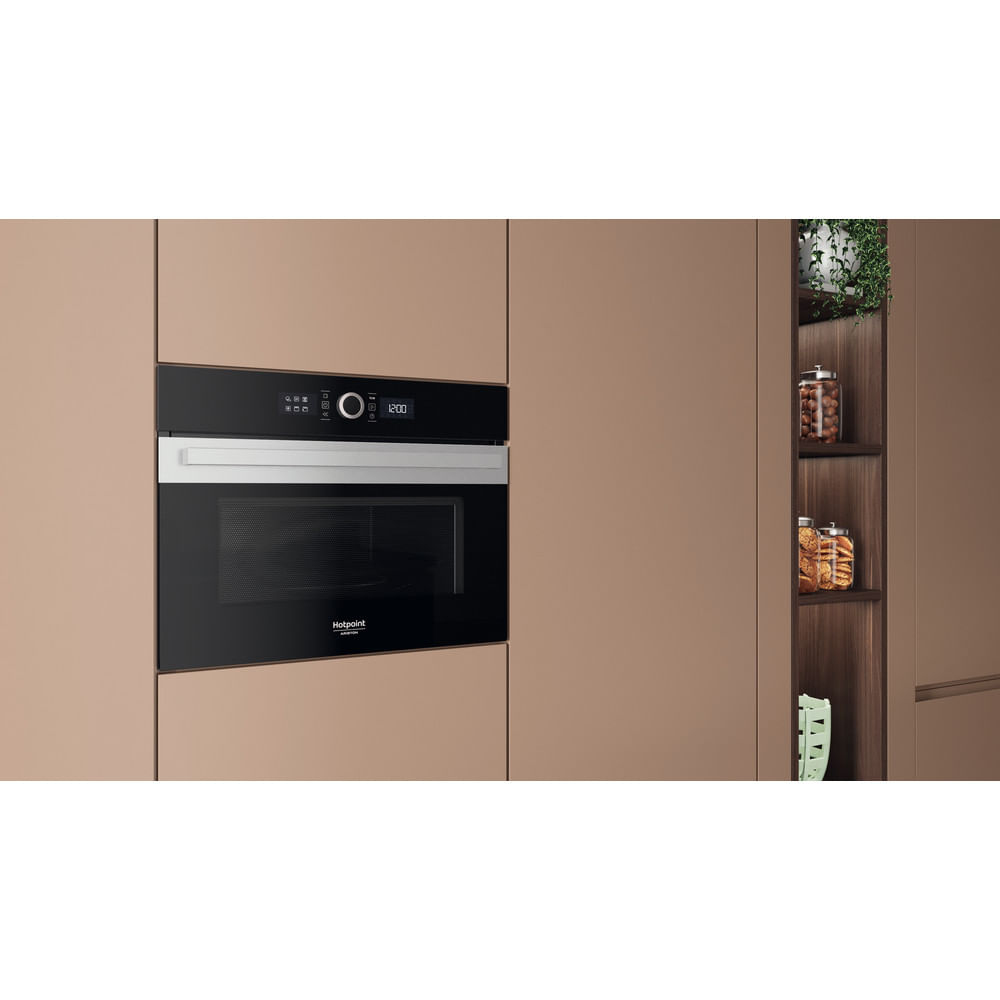 Forno a microonde nero a incasso con finiture argentate. Armadi sullo sfondo. Vasi con cibo e una pianta visibili a destra.