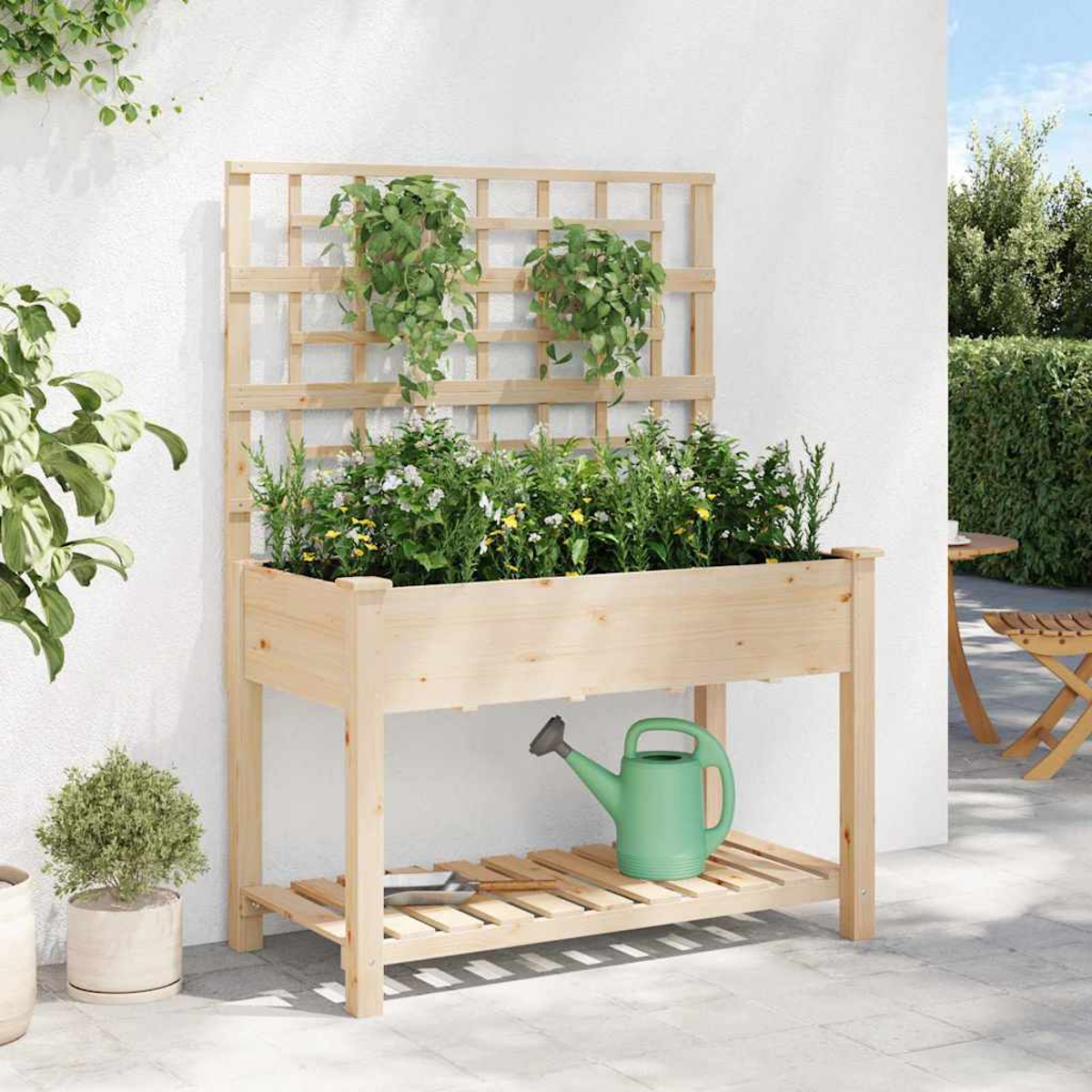 Fioriera in legno con piante, traliccio e un annaffiatoio verde su un patio. Piante verdi sono nella fioriera e appese al traliccio.