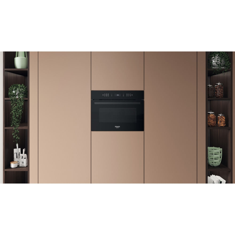 Un forno a microonde nero è integrato in un mobile da cucina beige. Ripiani su entrambi i lati mostrano piante, barattoli e oggetti da cucina.